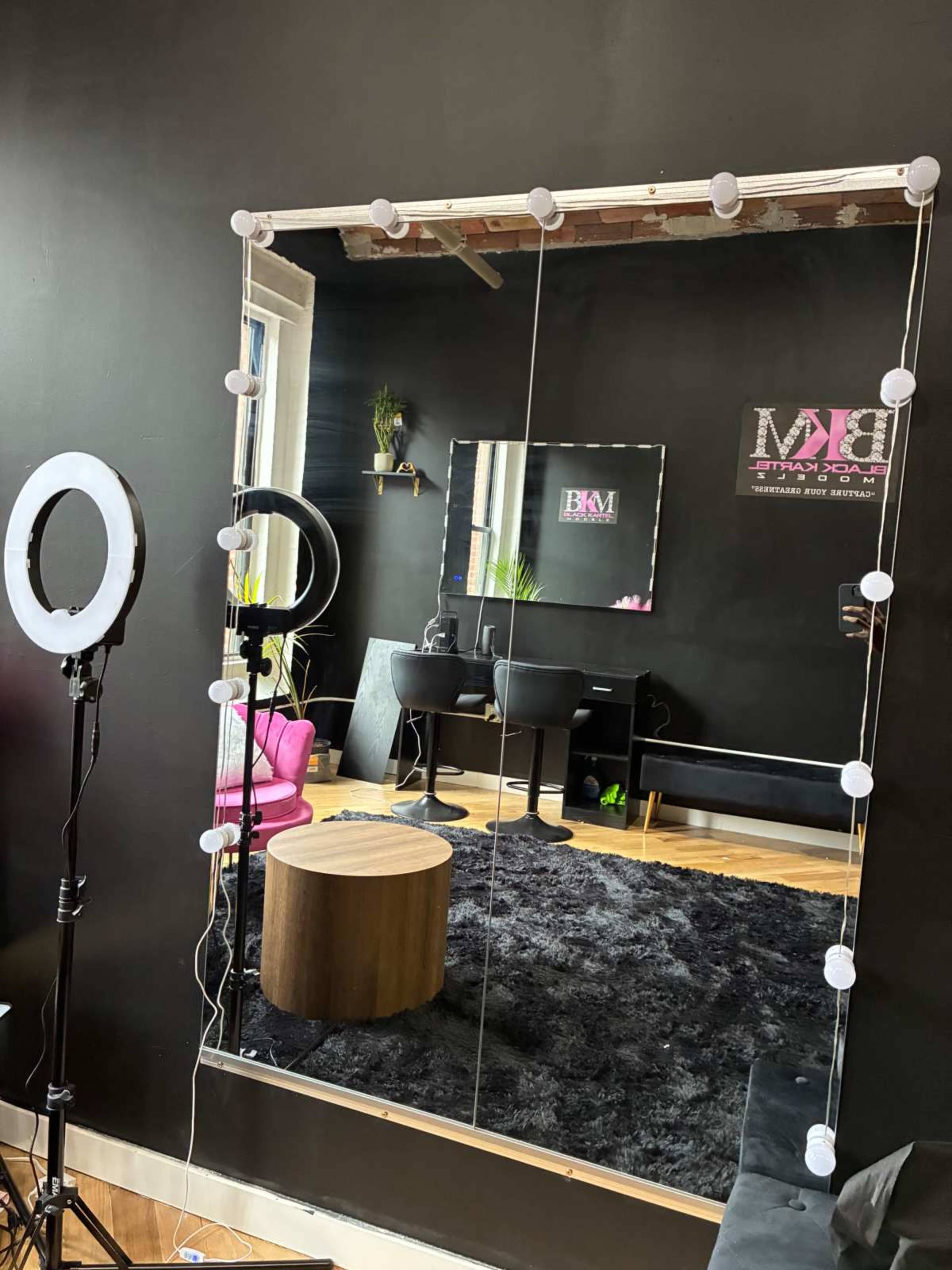 The image shows a modern beauty salon with a large mirror framed with lights, a round wooden table, and seats in a black and pink decor.