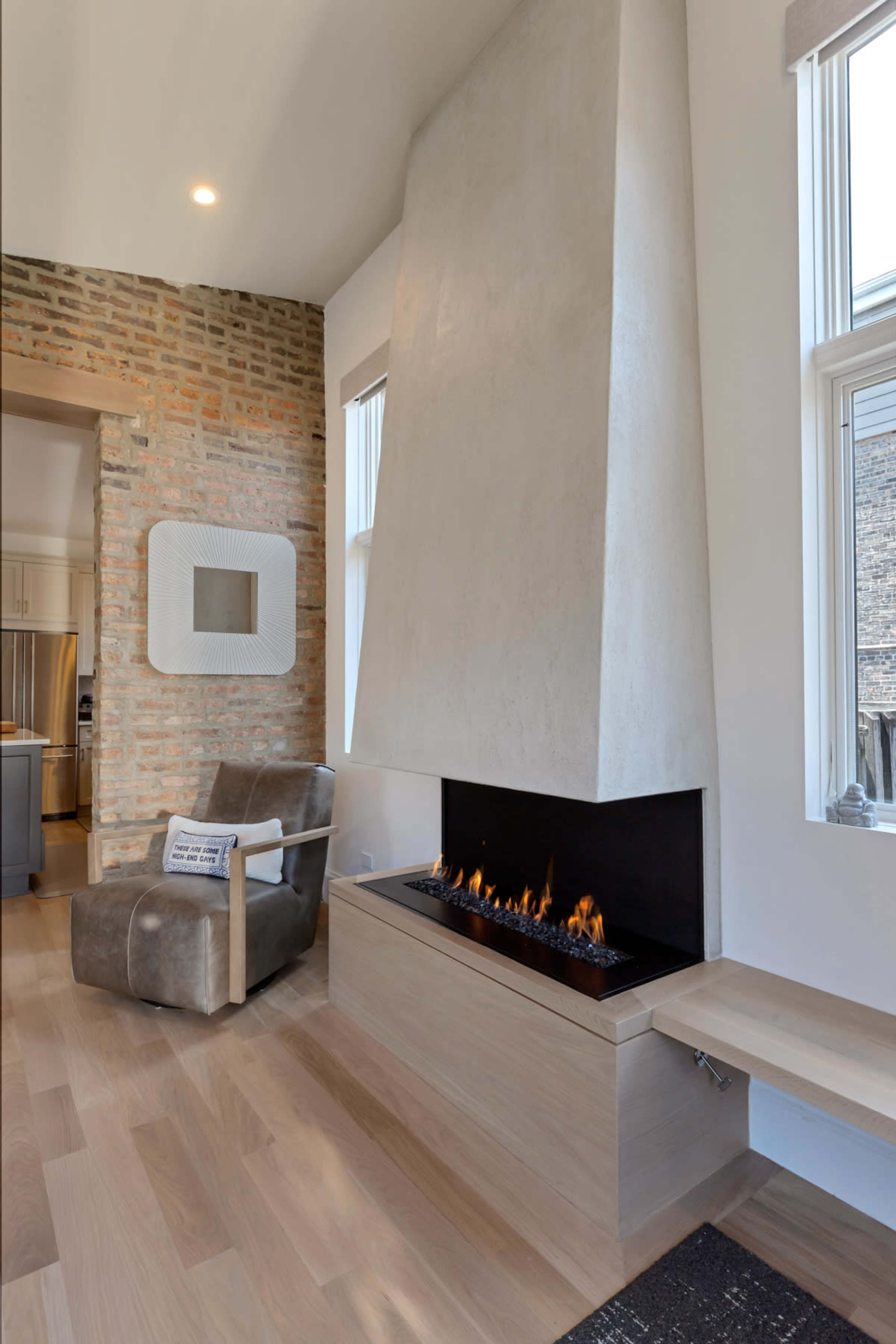 The image shows a modern living space featuring a tall, angled concrete fireplace with a linear gas flame, surrounded by a mix of brick and smooth white walls, complemented by a wooden bench and a cozy armchair.