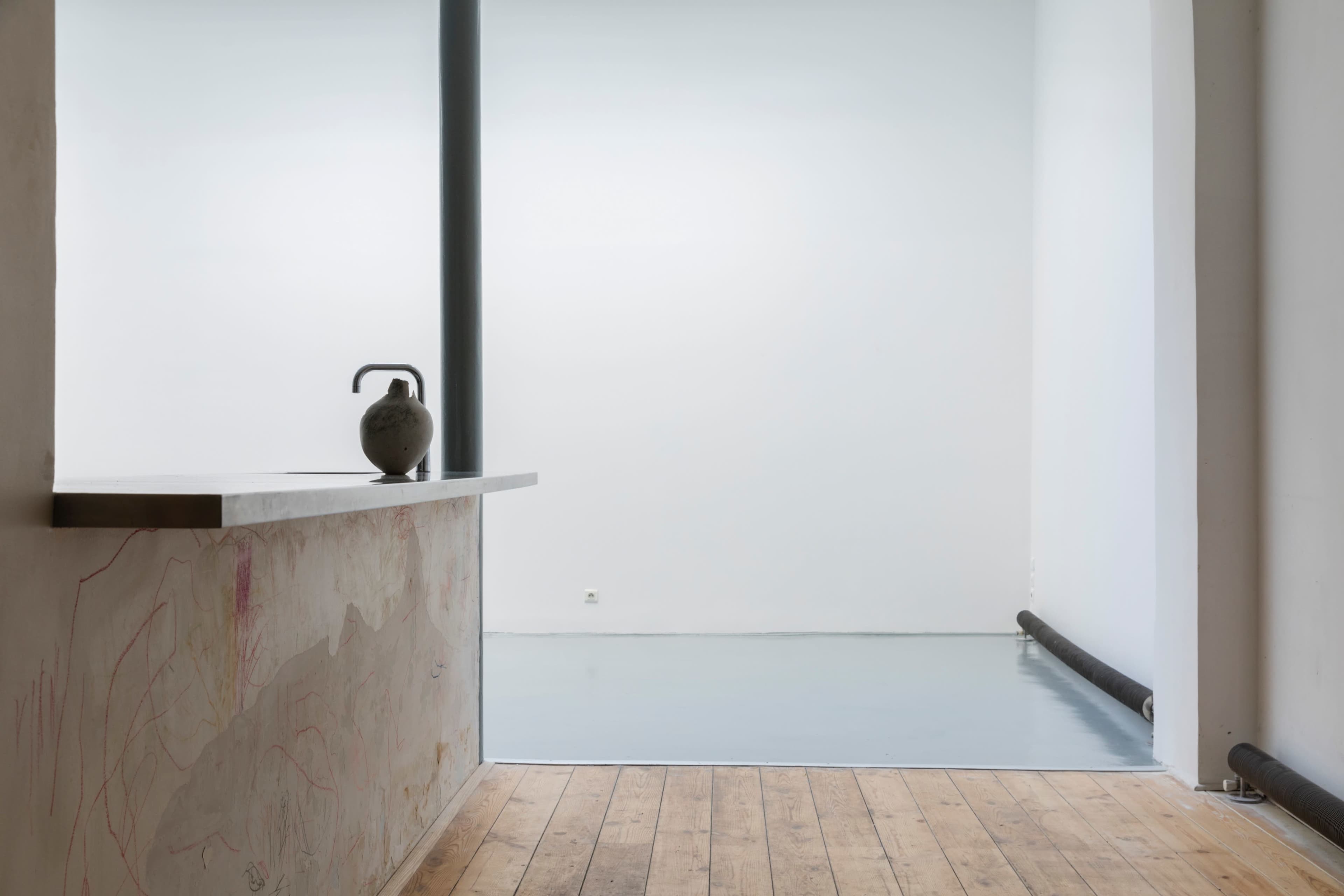 An empty interior space features a minimalist countertop with a vase, plain white walls, and wooden flooring.
