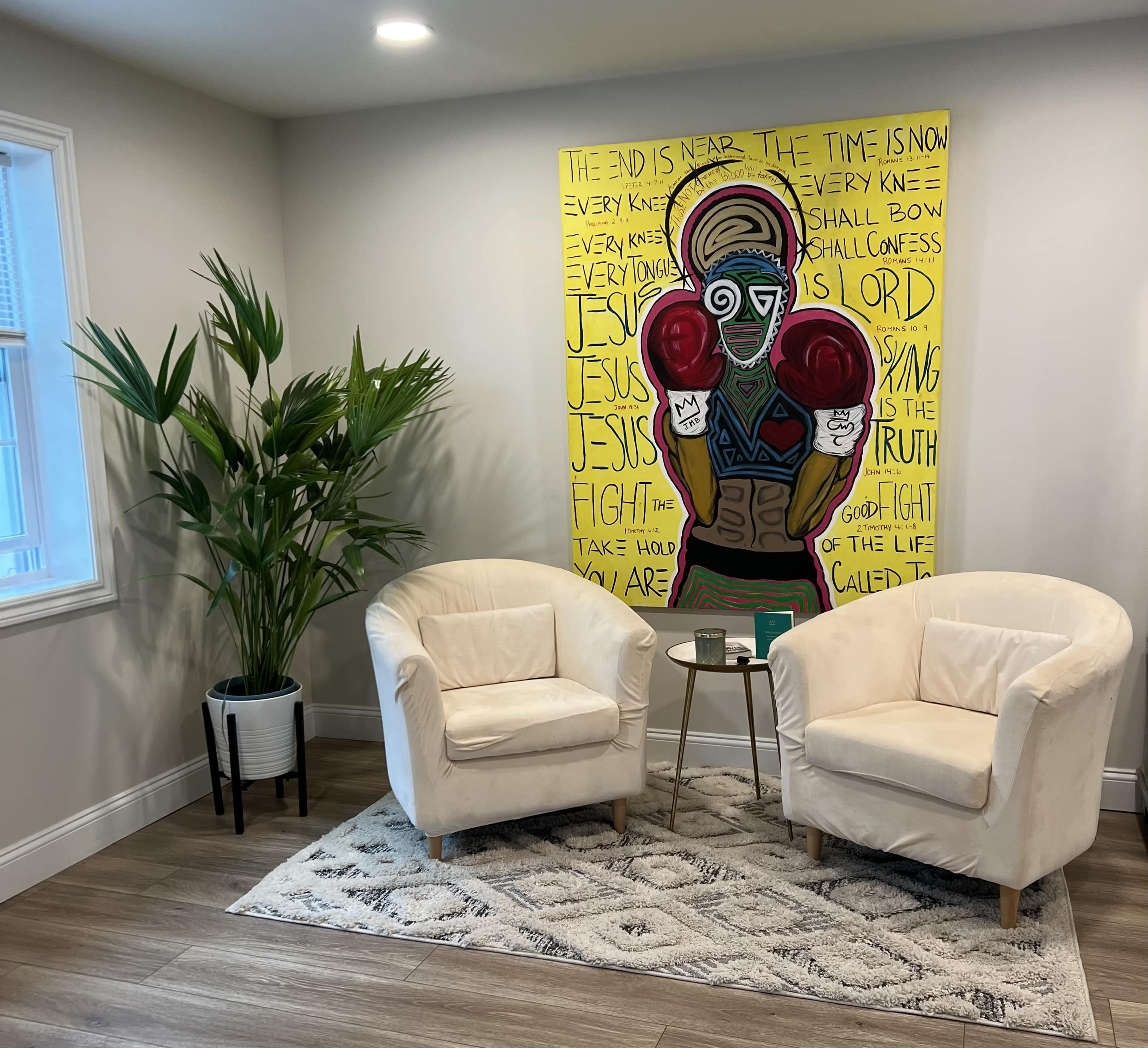 A bright room features two white chairs flanking a small side table, set against a vibrant mural with bold text and imagery on the wall.