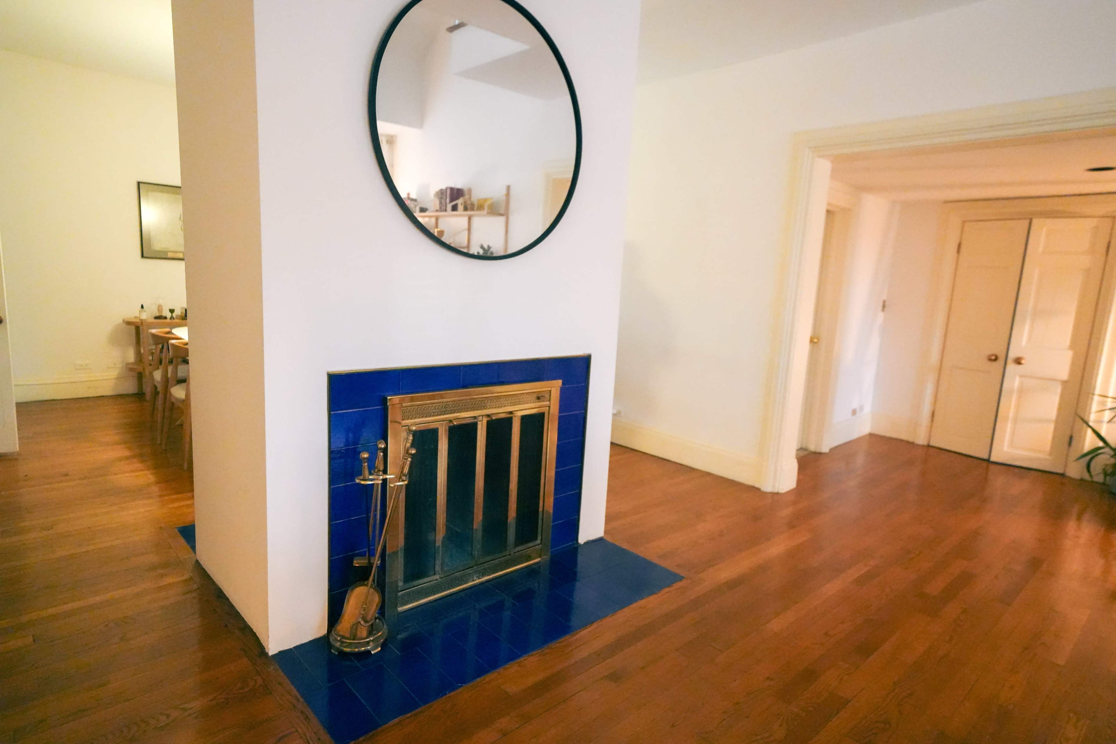 A living space featuring a fireplace with a blue tile surround and a circular mirror above it, with hardwood floors and doorways leading to other rooms.