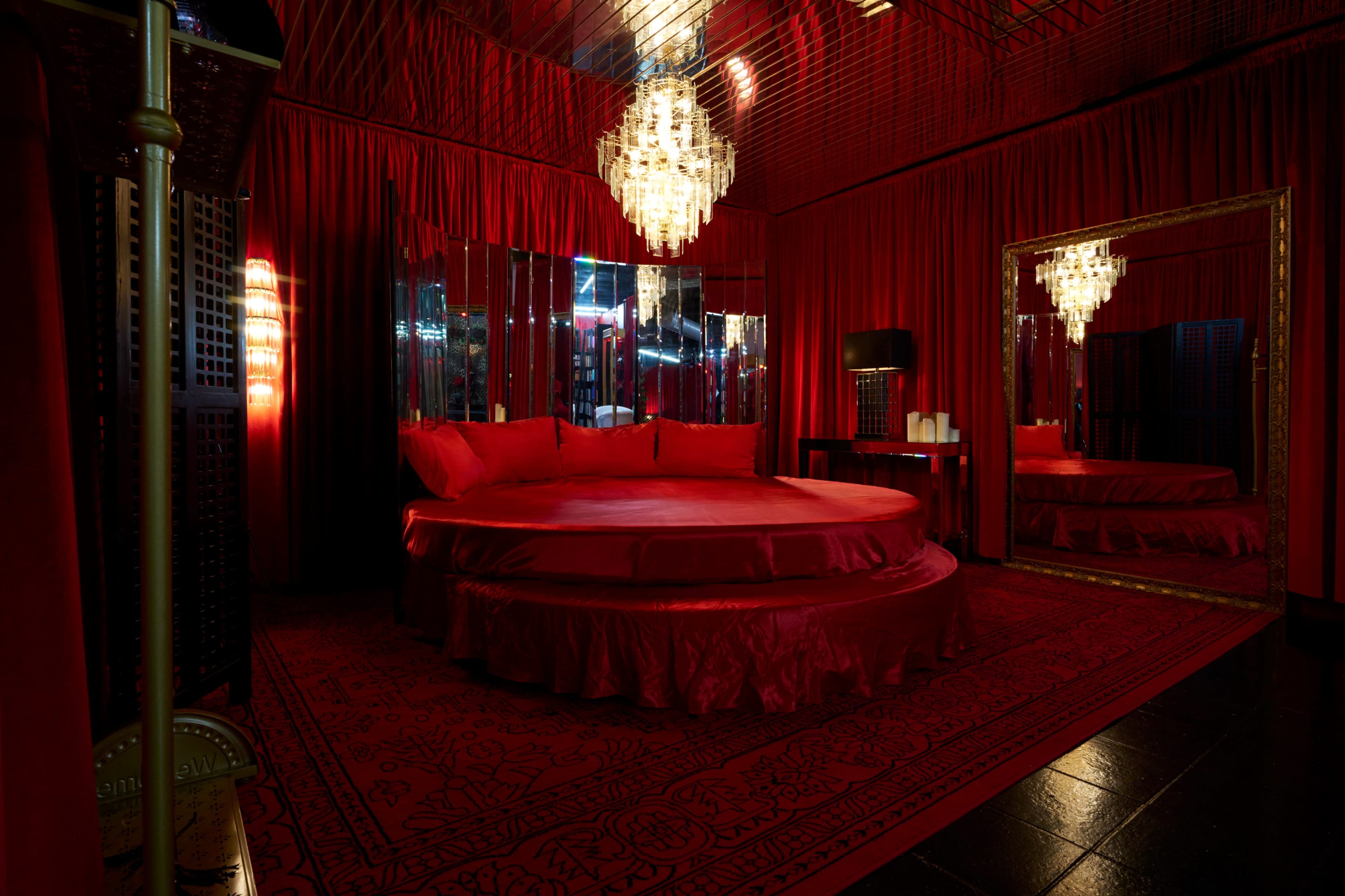 A circular bed surrounded by red drapes and walls, illuminated by a crystal chandelier and mirrored surfaces.
