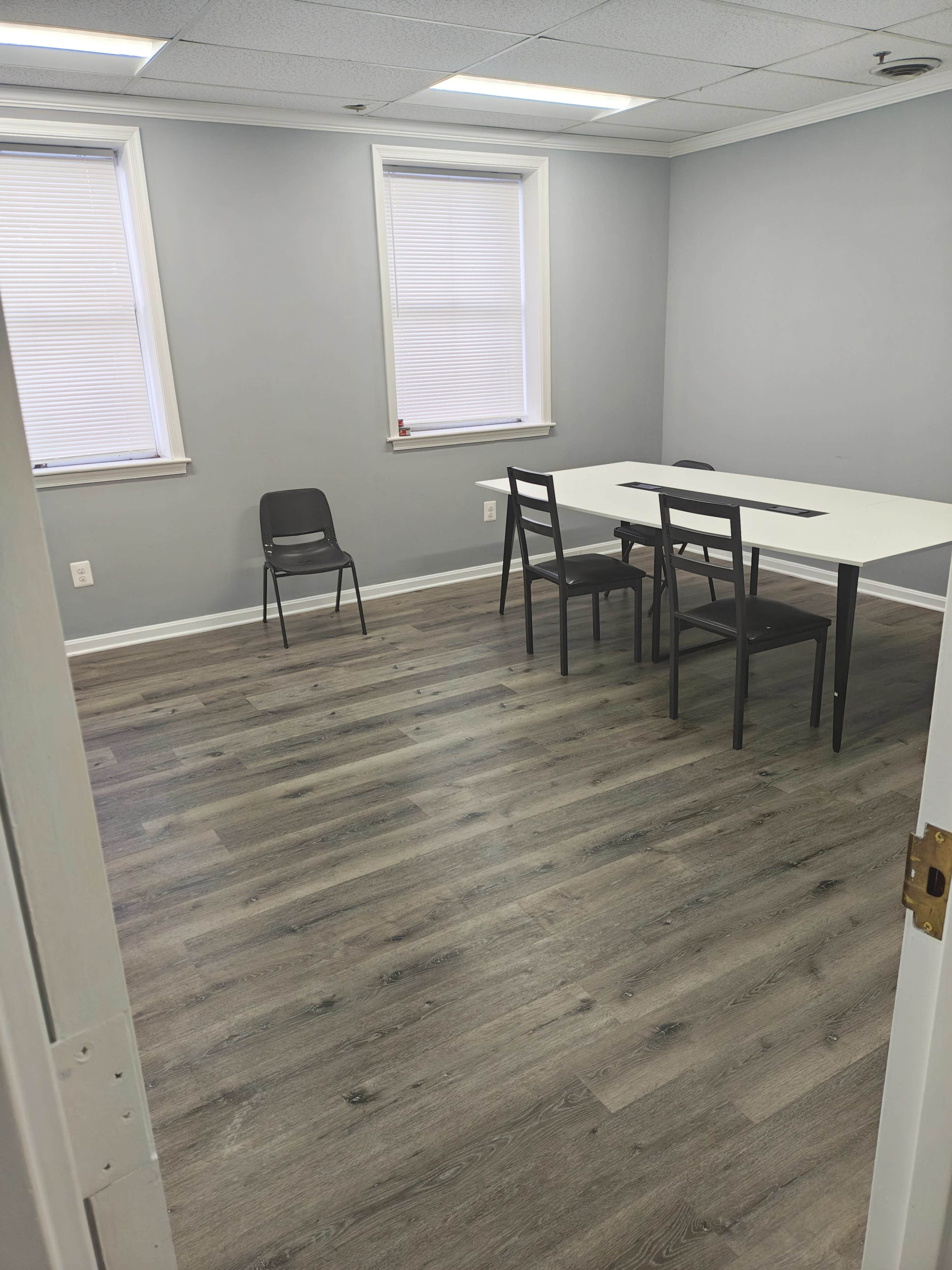 The image shows a small meeting room with a white table and four black chairs, featuring gray walls and laminate flooring.