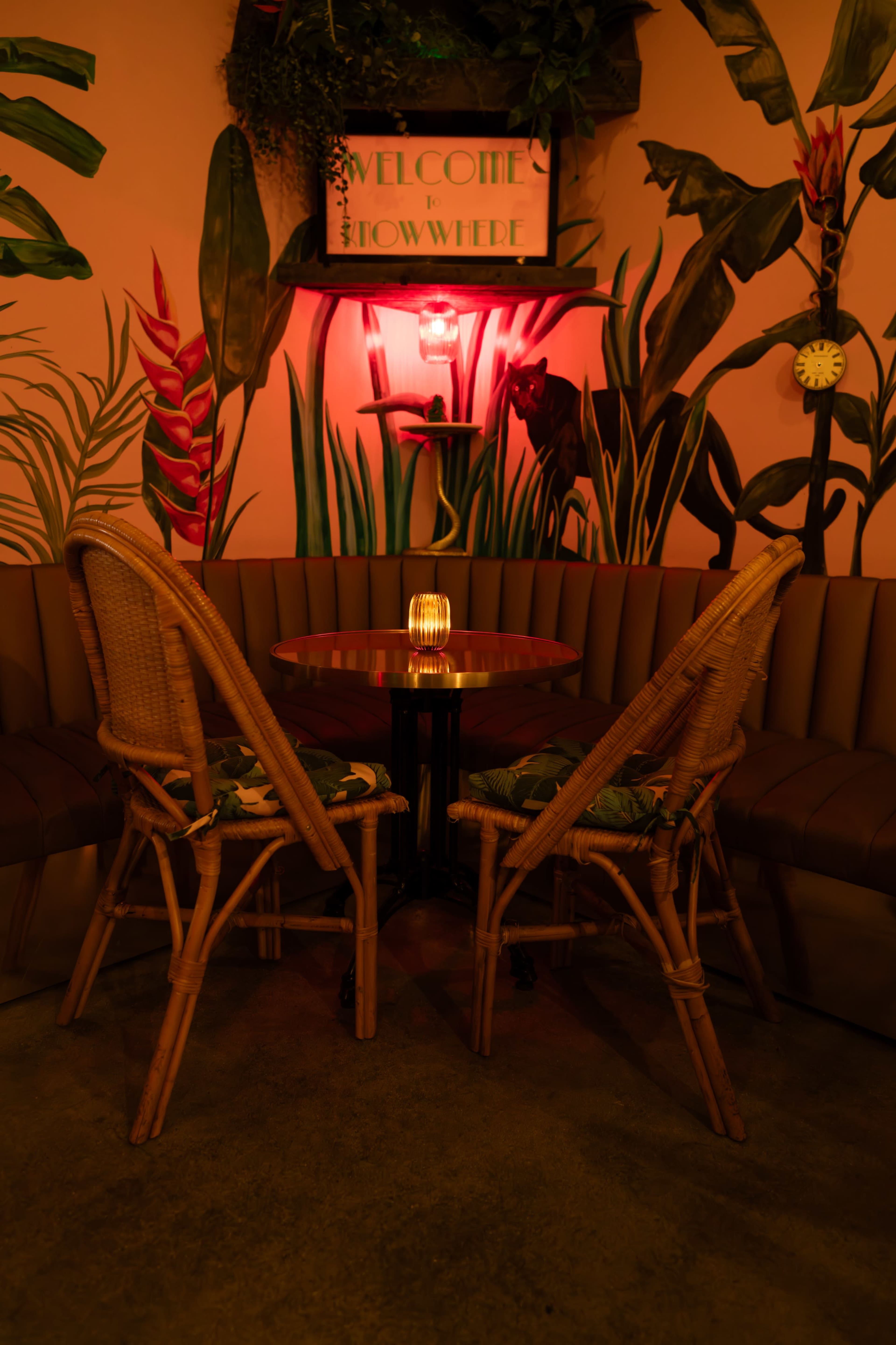 A cozy seating area features a round table illuminated by a warm light, flanked by two rattan chairs against a backdrop of vibrant tropical plants painted on the wall.