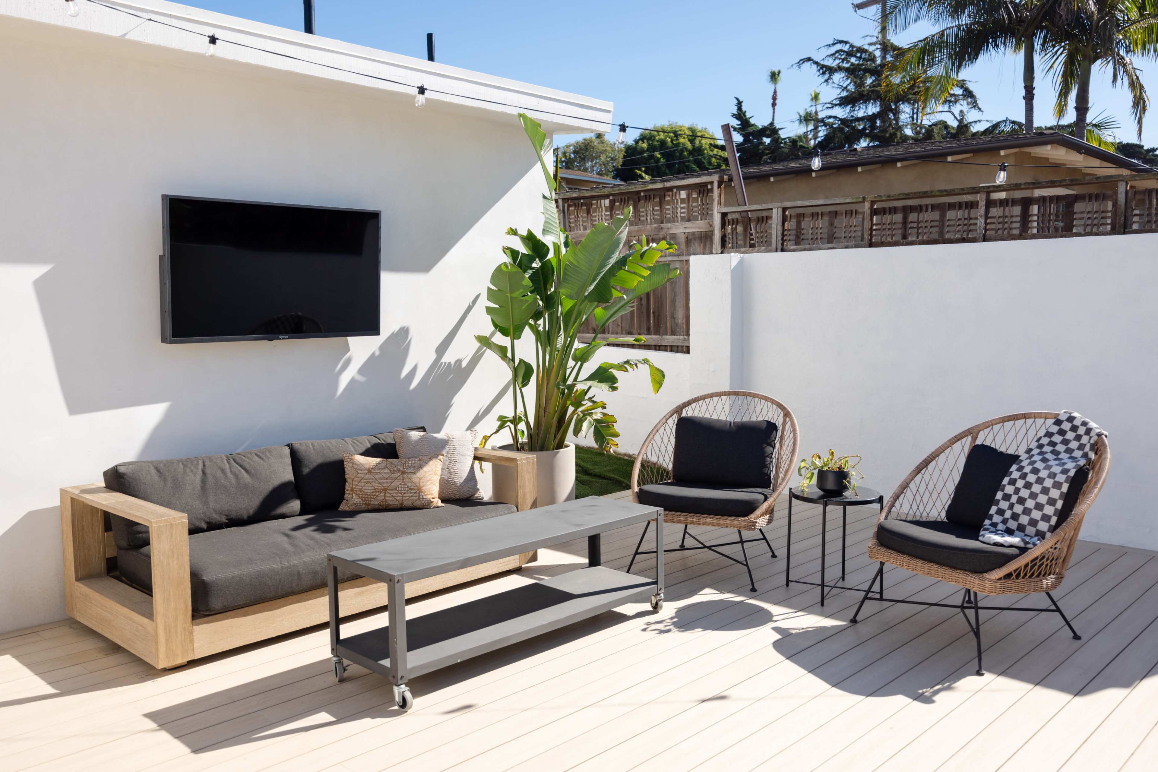 The image shows a modern outdoor seating area featuring a gray sofa, two rattan chairs, and a coffee table on a wooden deck, with a television mounted on the wall and potted plants nearby.