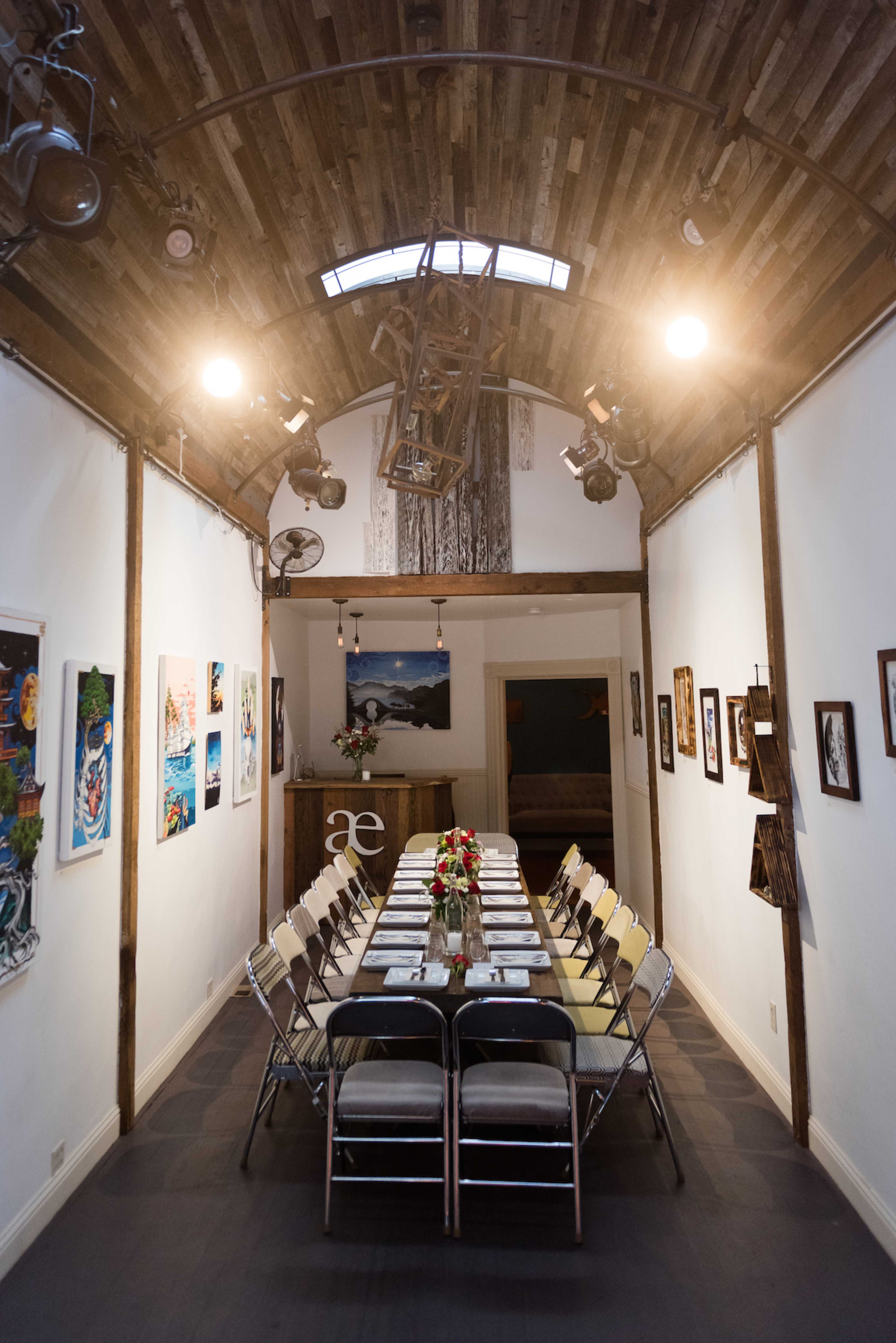 A long table is set for a meal in a well-lit room featuring wooden beams and artwork on the walls.