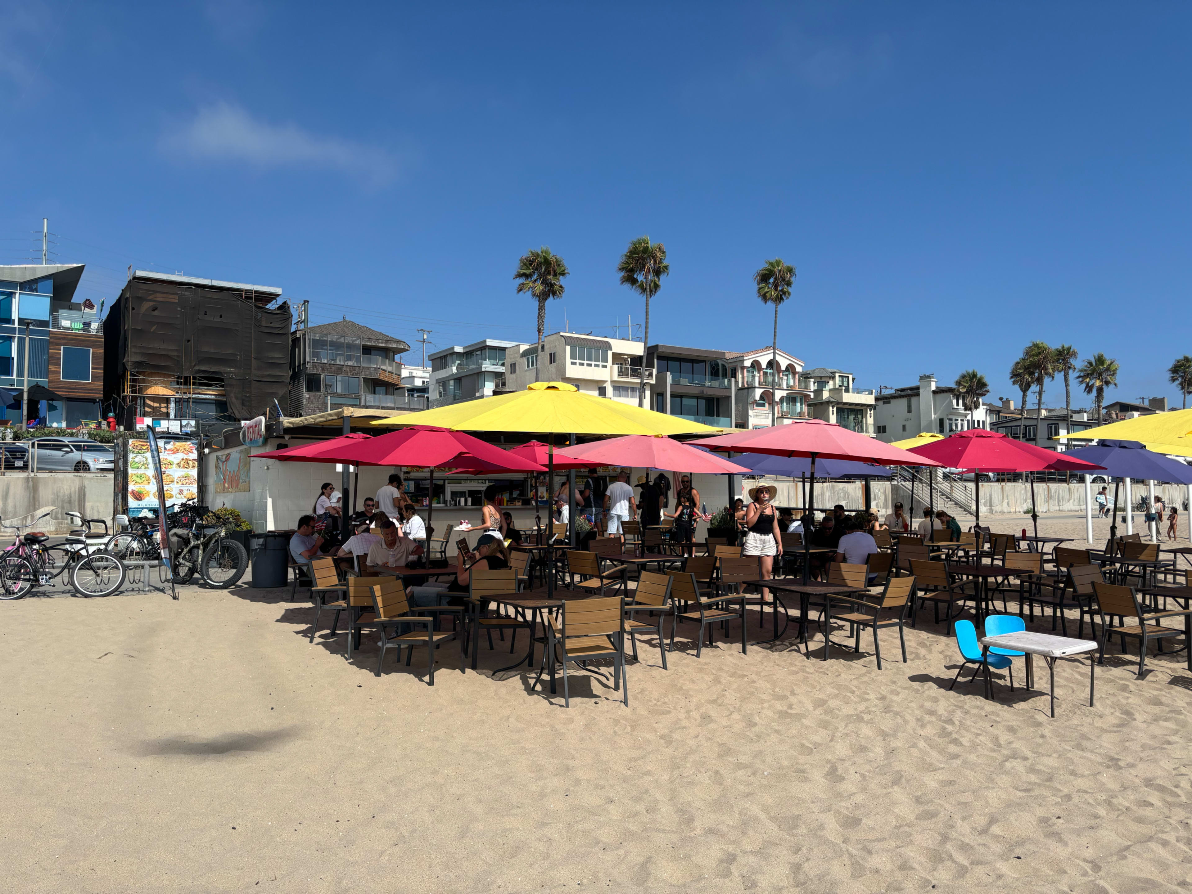 A beachside dining area features colorful umbrellas over tables with patrons enjoying the view and sand in the background.