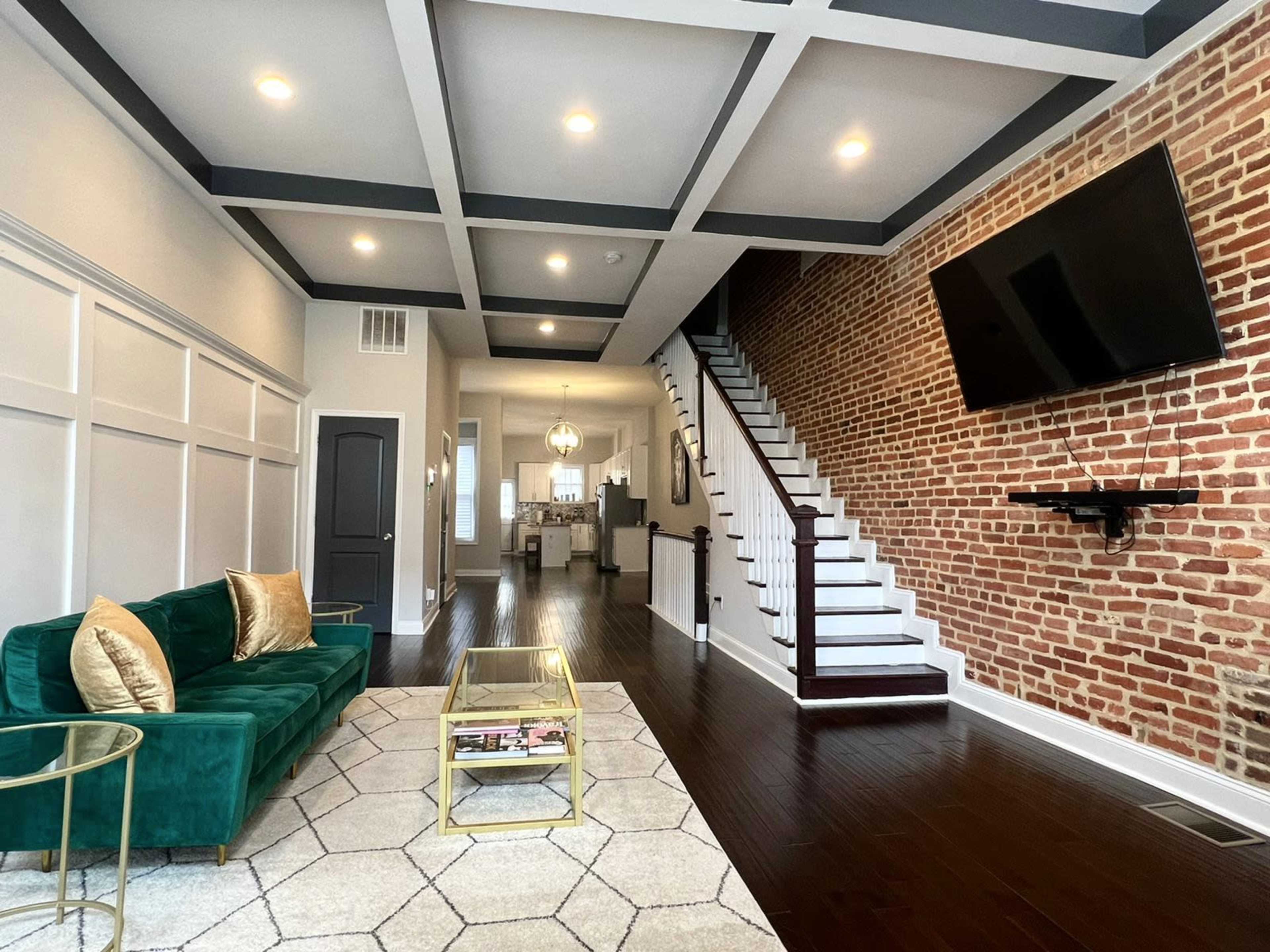 A modern living room with a green sofa, a geometric rug, a glass coffee table, exposed brick walls, and a staircase leading to an upper level.