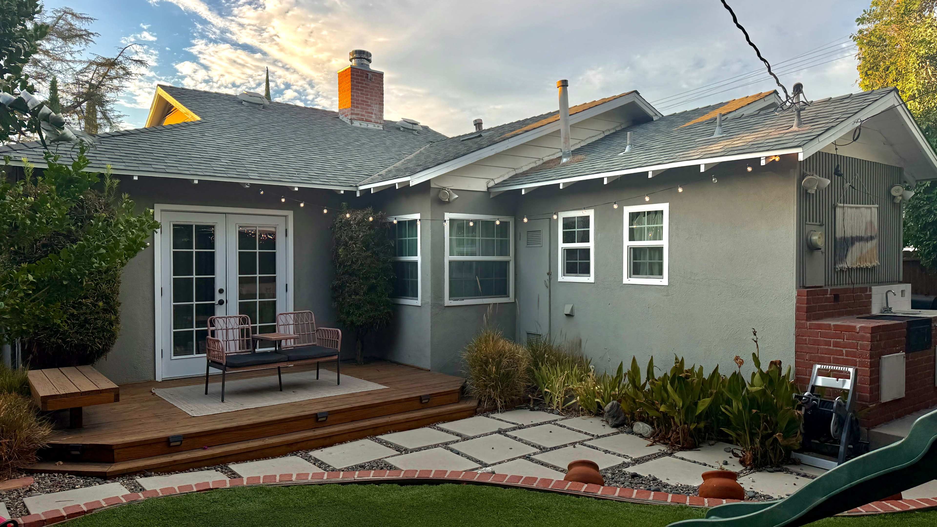 The image shows a small backyard with a wooden deck, two chairs, and a landscaped area featuring plants and stone pathways, adjacent to a gray house with multiple windows.