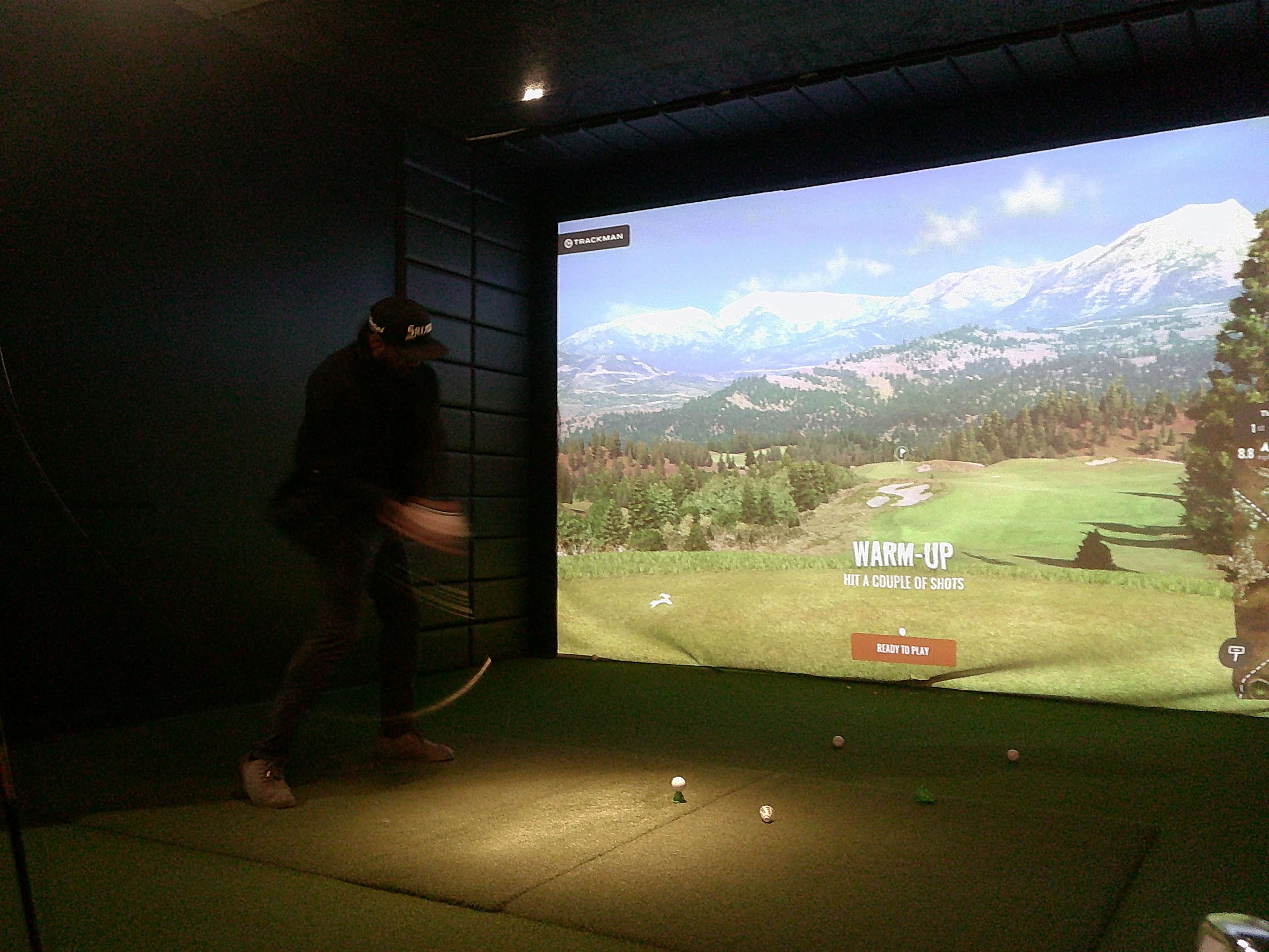 A golfer is practicing their swing in an indoor simulator that displays a scenic golf course on a large screen.