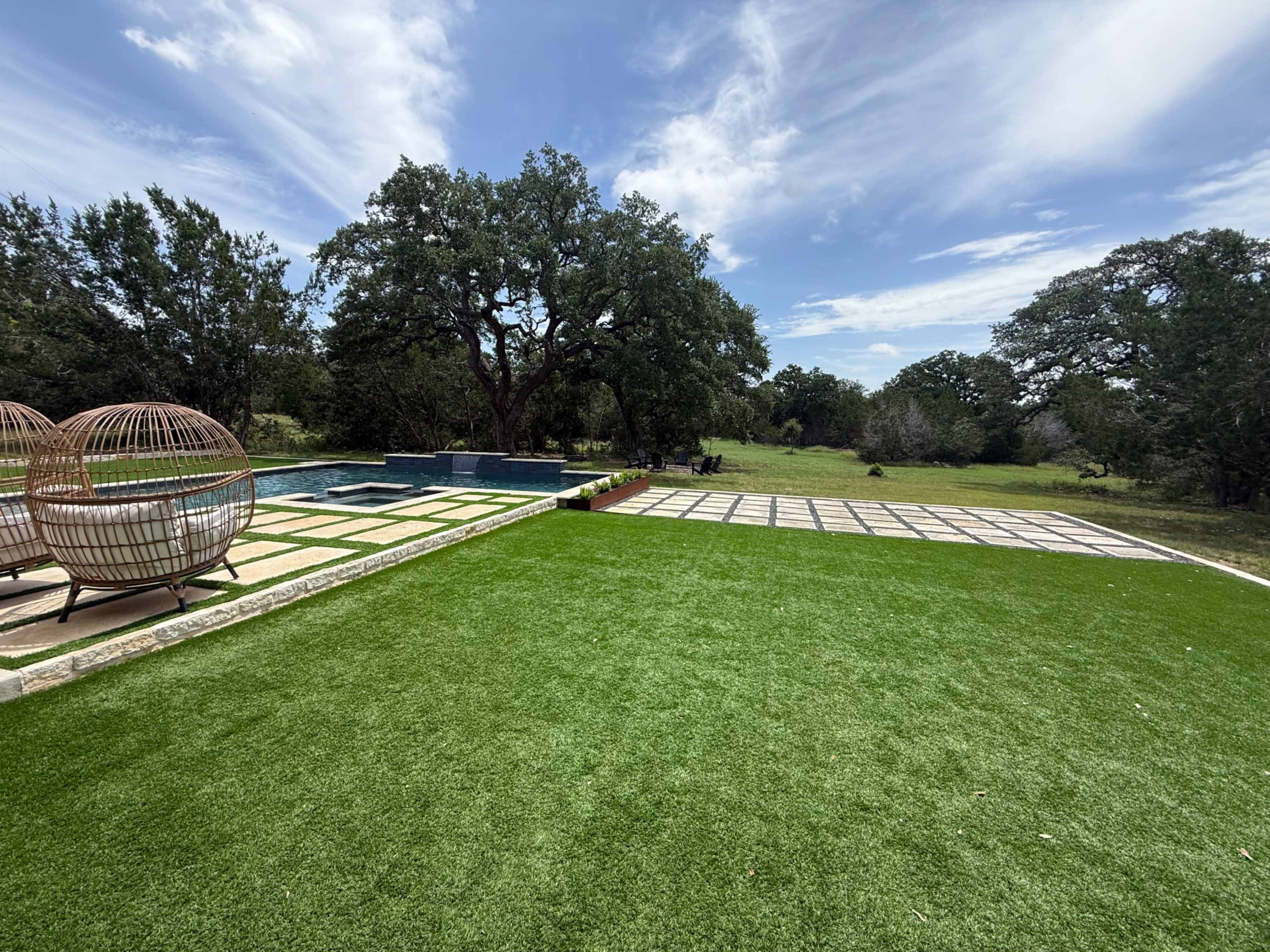 A backyard features a swimming pool, a patio with a tiled surface, and a large expanse of artificial grass, surrounded by trees and open land.