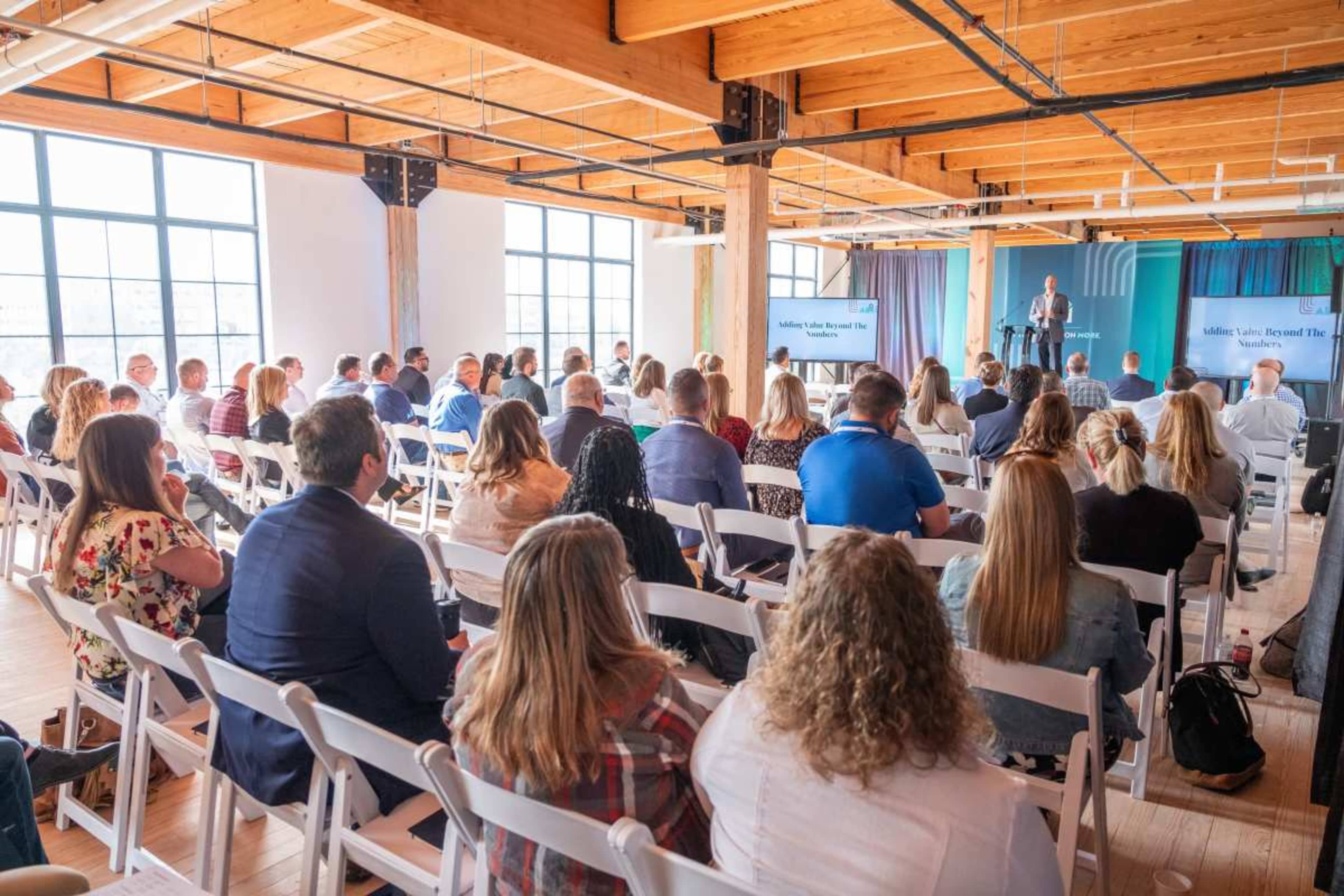 A group of people is seated in a large room with wooden beams, listening to a speaker presenting on a stage.