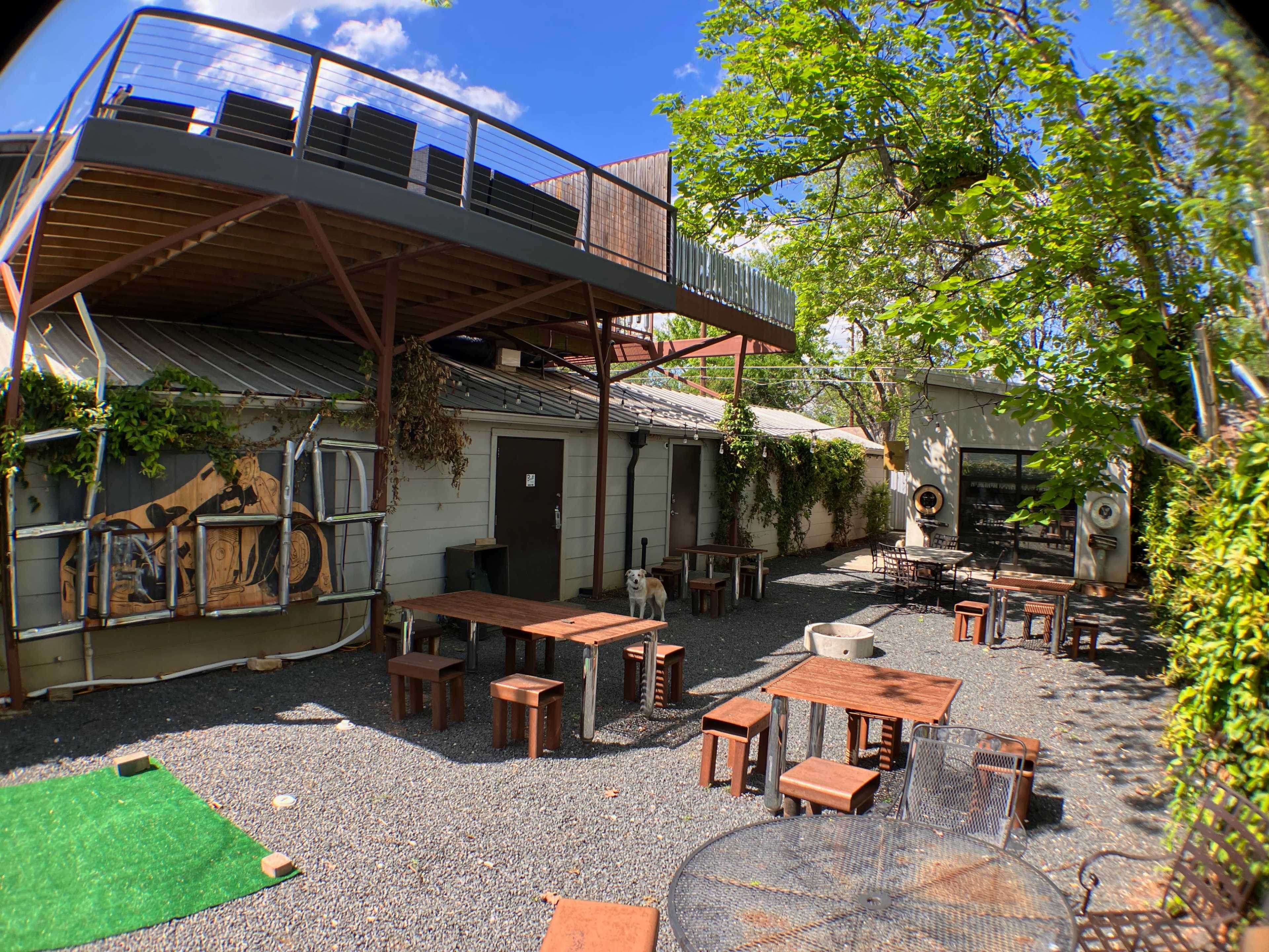 The image shows an outdoor area with gravel flooring, wooden tables and stools, a building with vines, and a second-story patio above.