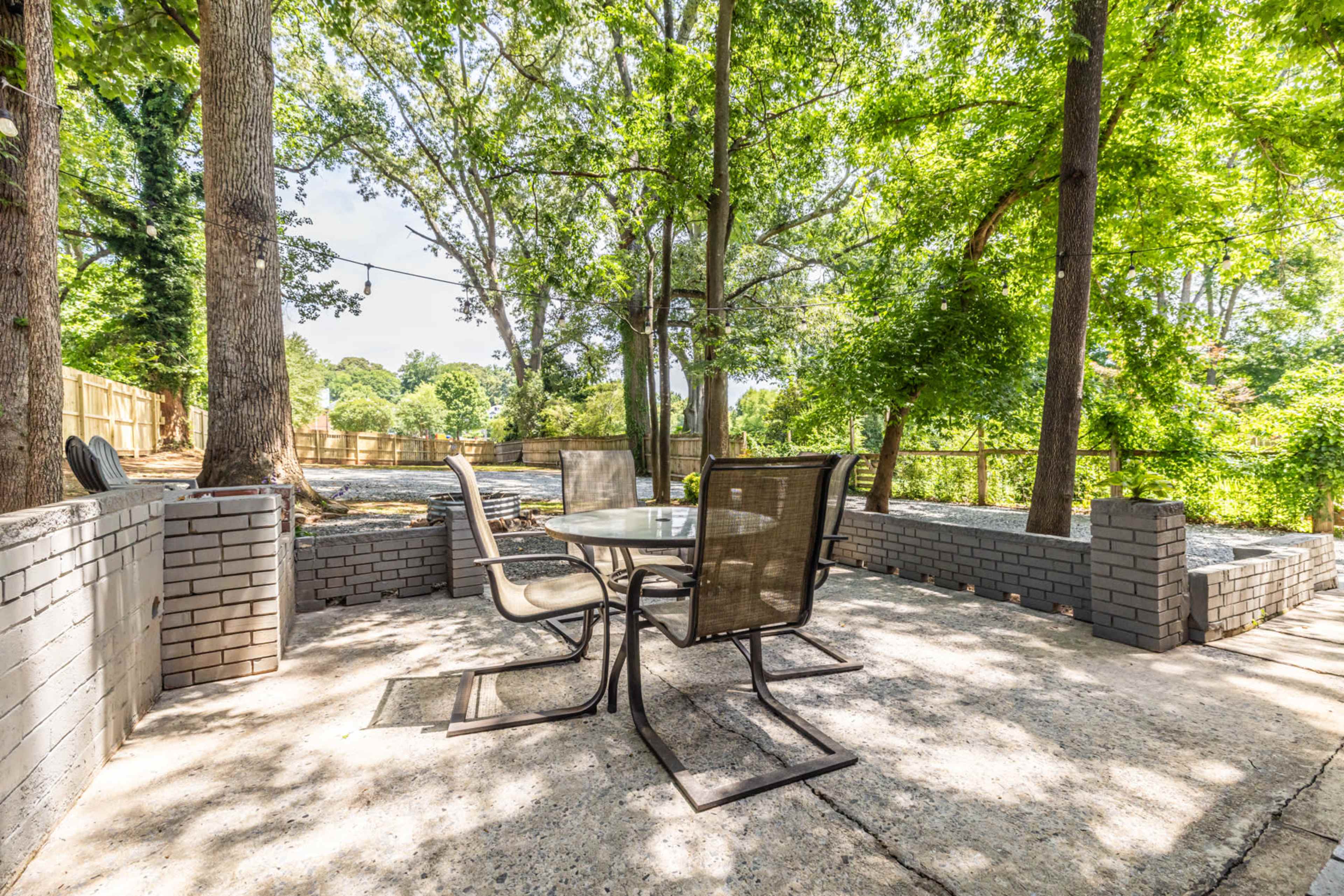 The scene features a patio area with a glass table and four chairs, surrounded by trees and a landscaped yard.