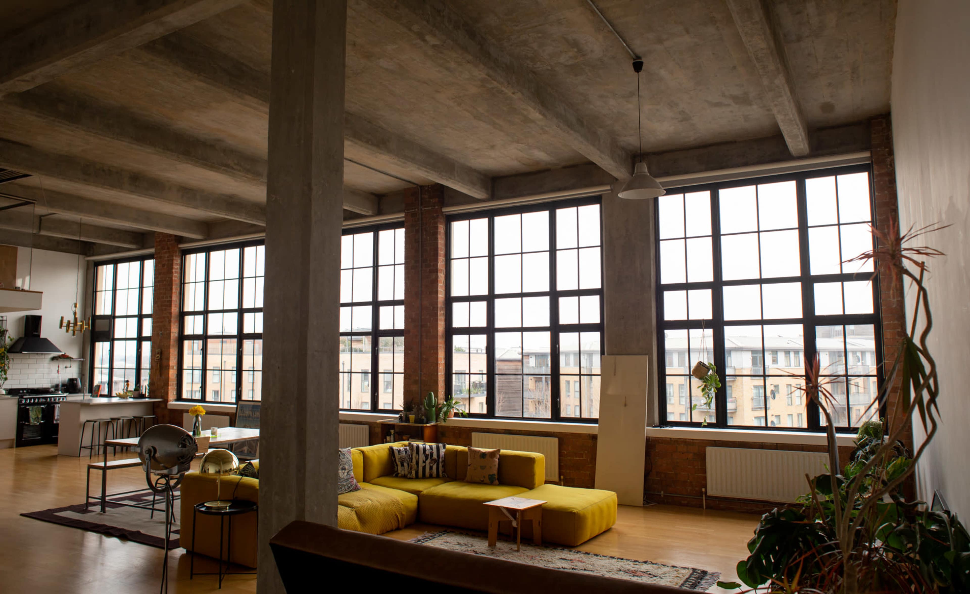 An industrial-style living room featuring large windows, a yellow sofa, and a wooden dining table.