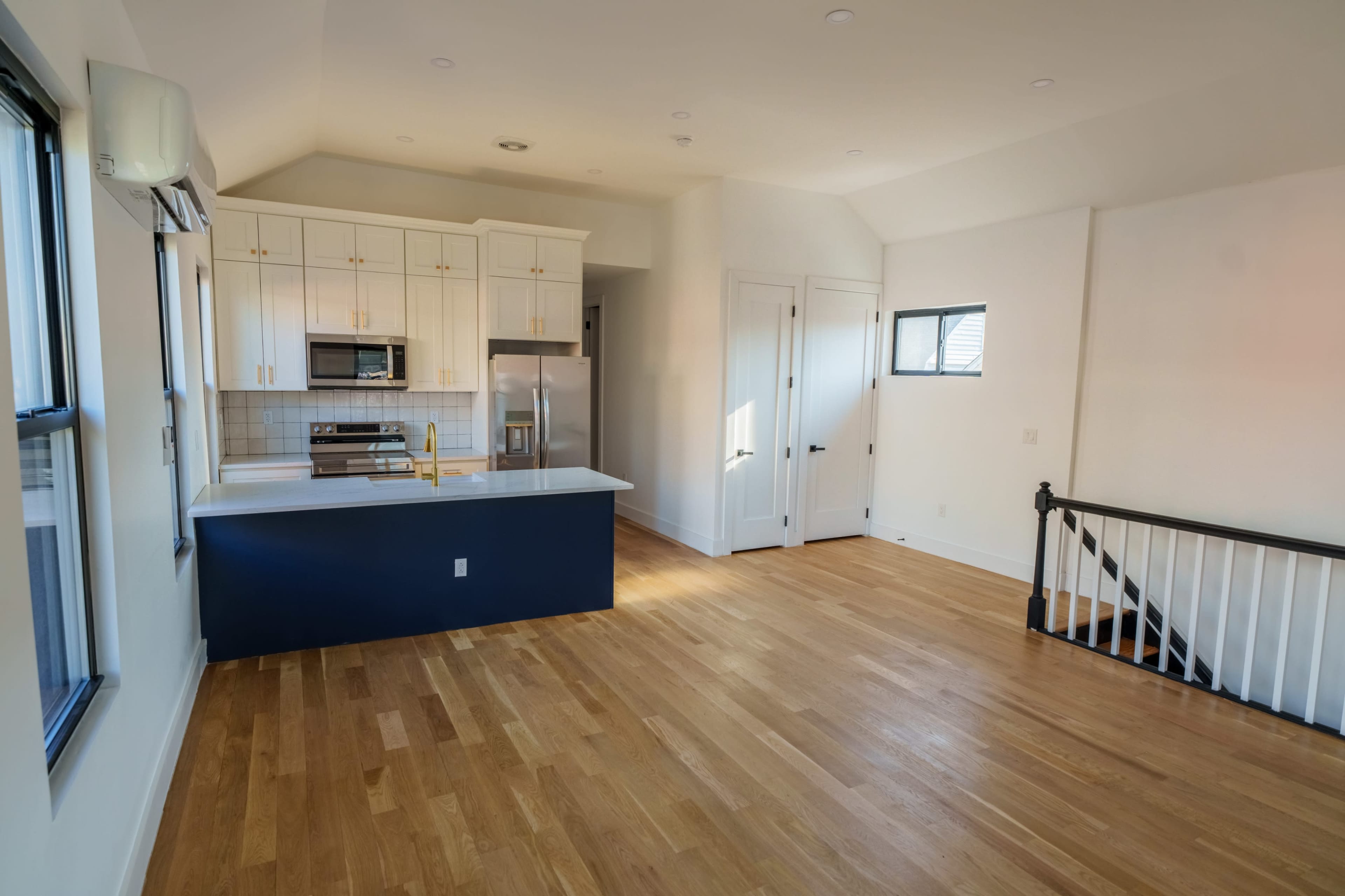 The image shows a small, modern kitchen with white cabinets and stainless steel appliances, adjacent to an open living area with hardwood floors and large windows.