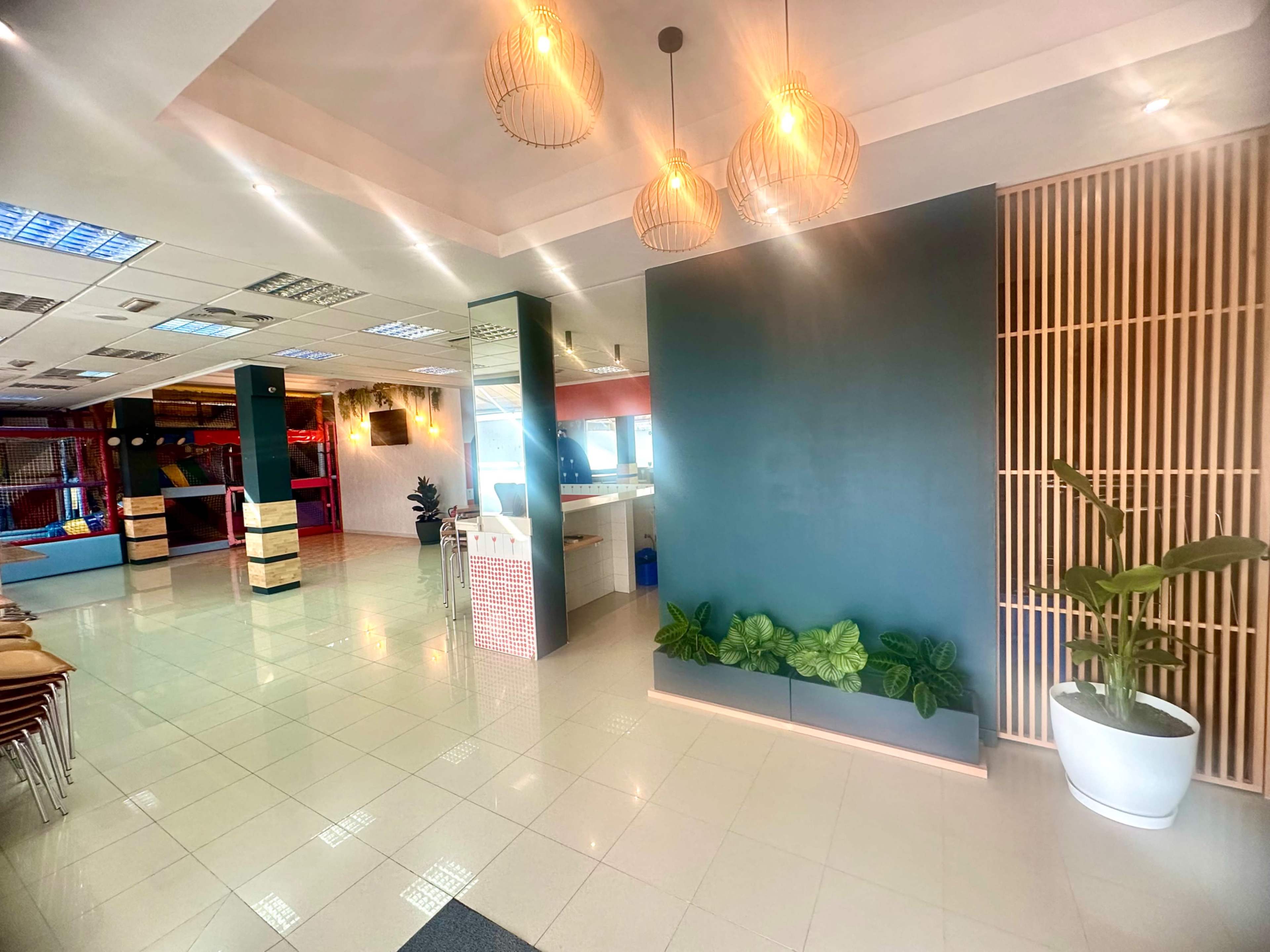 The image shows a spacious indoor area featuring a modern reception with pendant lighting, green plants along the wall, and an open layout that includes leisure and dining spaces.