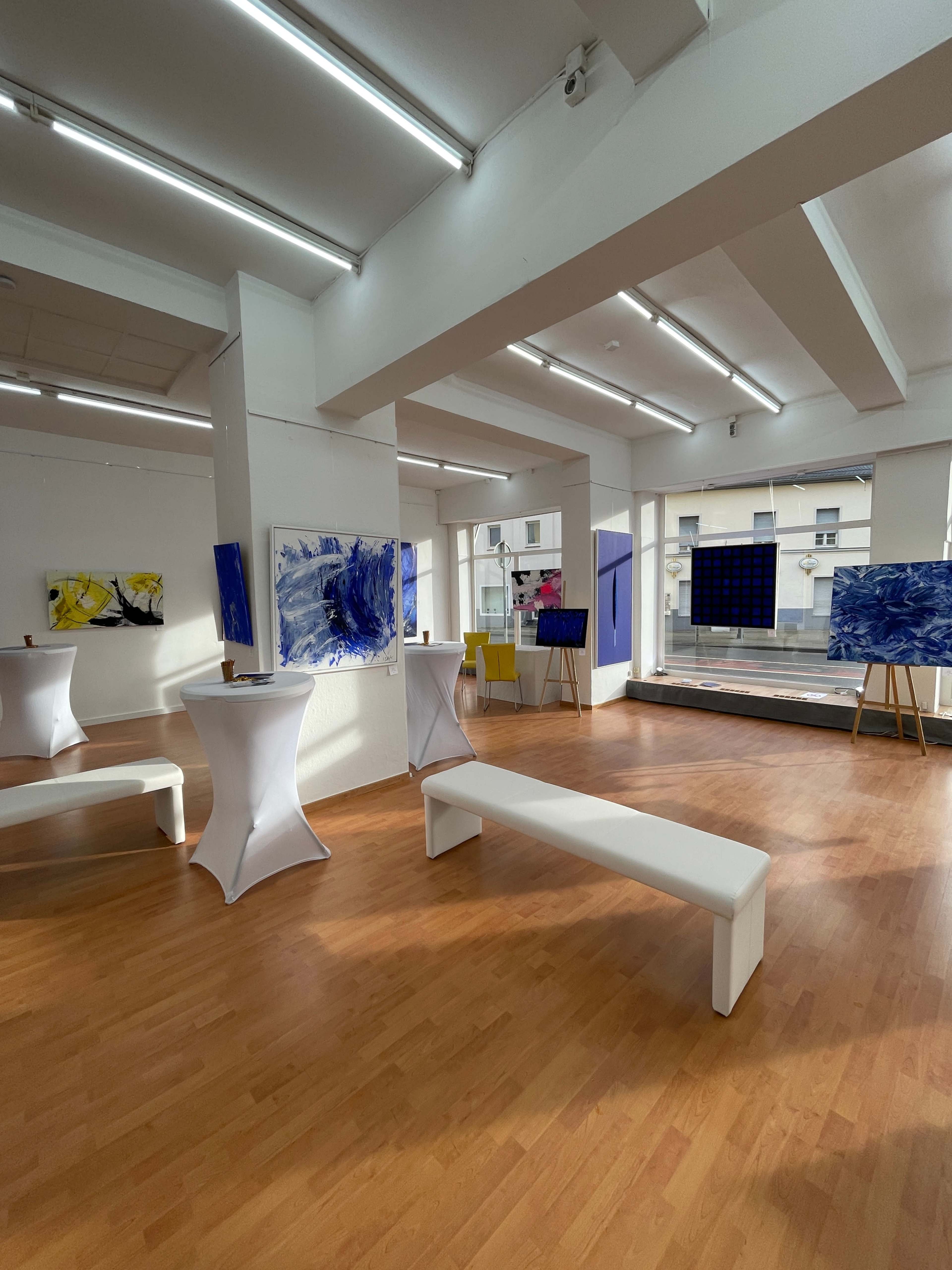 An art gallery featuring abstract paintings on the walls, with white benches and table setups arranged in the space.