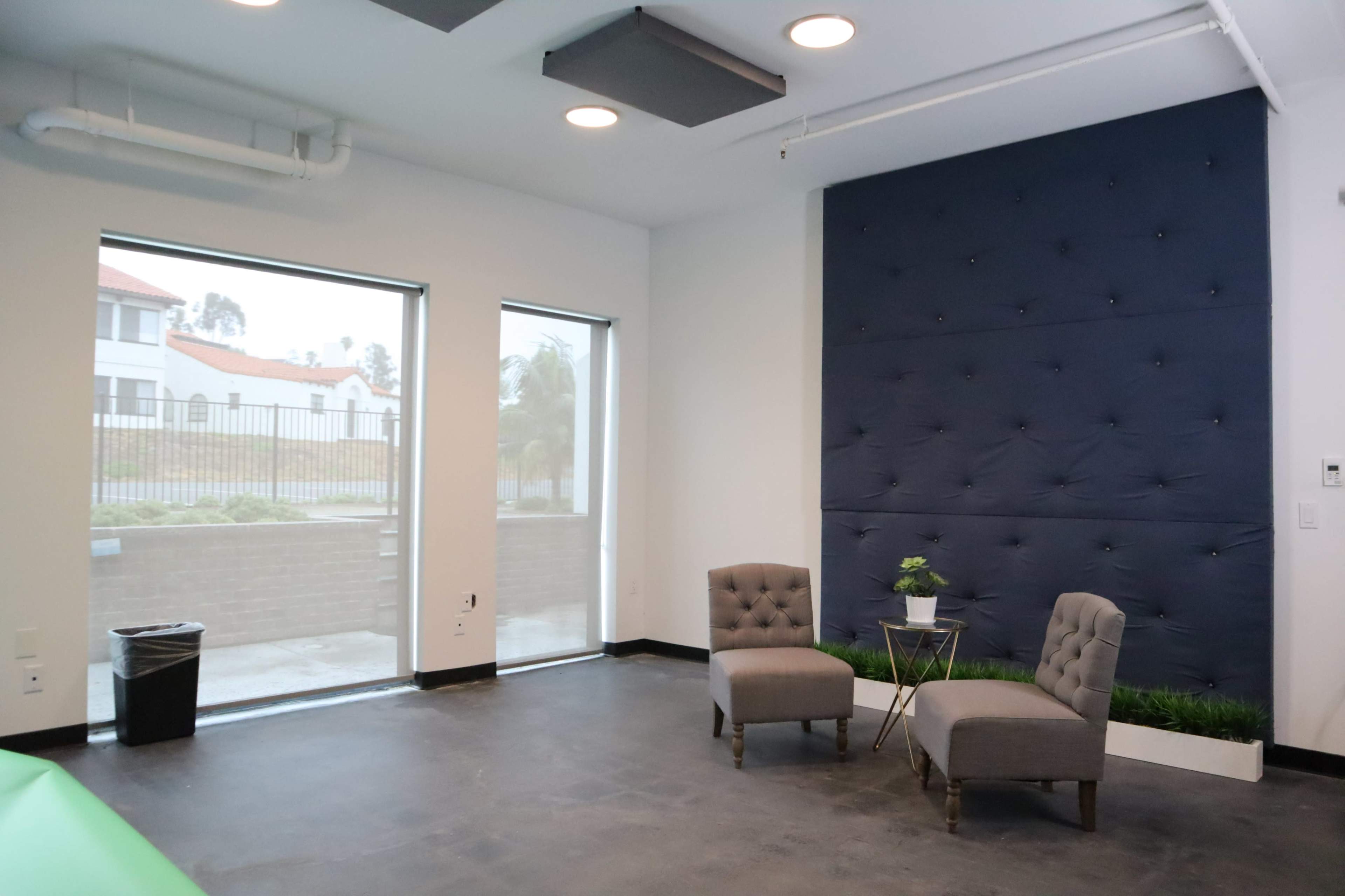 A modern room with two gray armchairs facing a small table against a blue tufted wall and large windows revealing a view of an outside area.