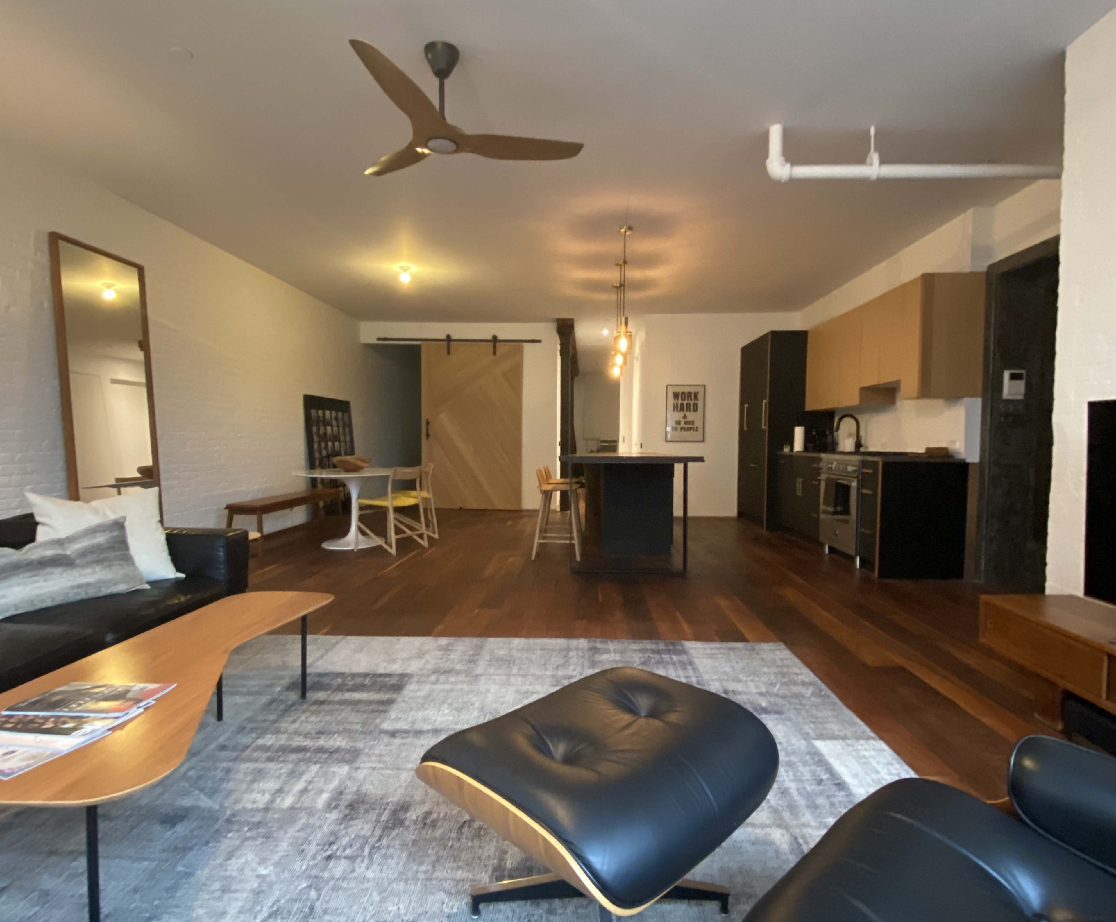 A modern living space featuring hardwood floors, a sectional sofa, a dining area, and a kitchen with wooden cabinetry.
