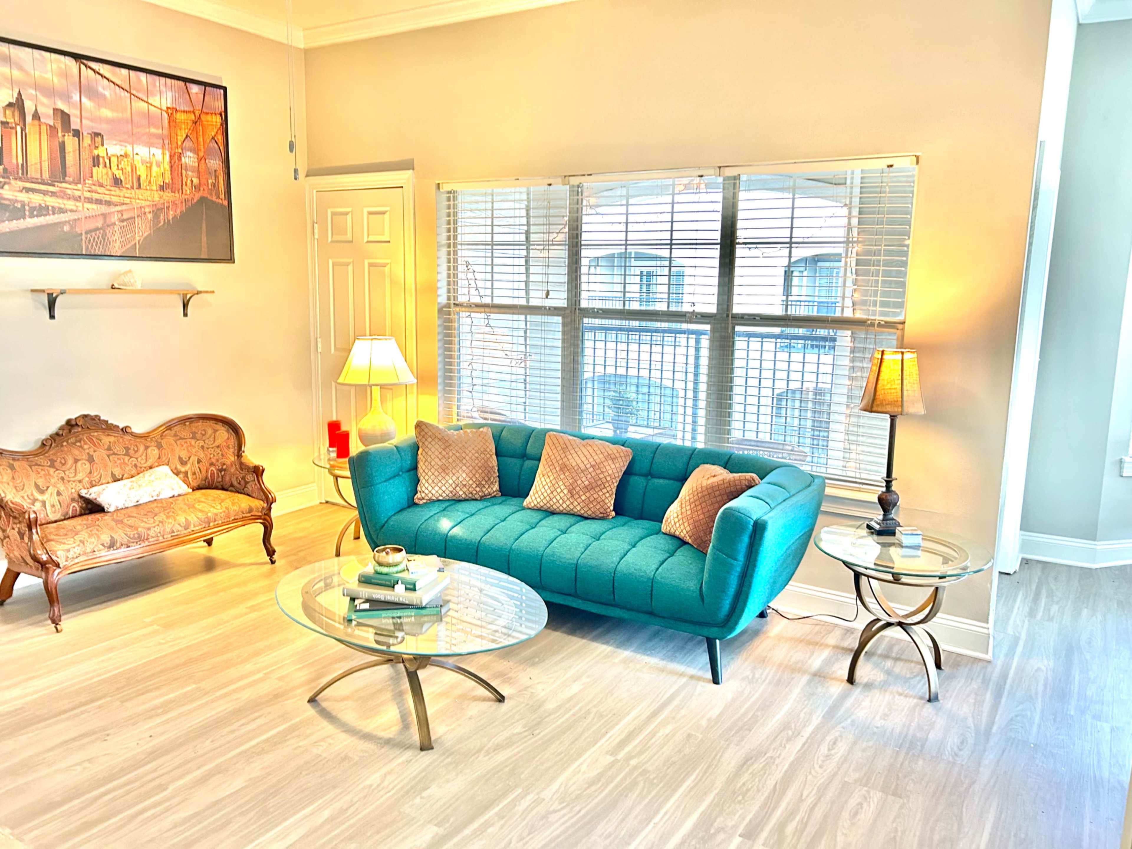The image shows a bright and modern living room featuring a teal couch, a vintage settee, glass coffee tables, and lamps illuminating the space.