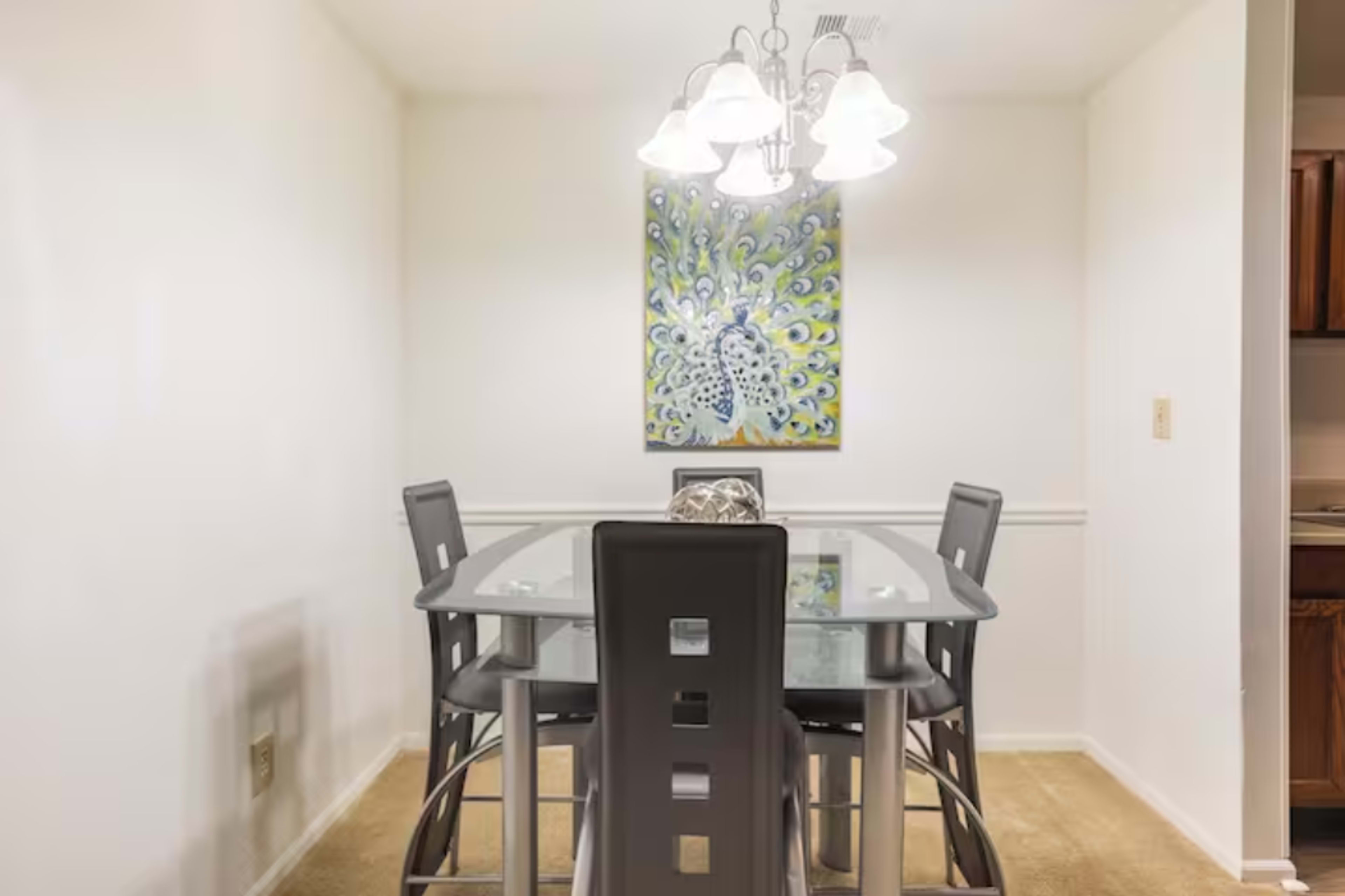 A glass dining table with four black chairs is illuminated by a pendant light, while a colorful artwork hangs on the wall.