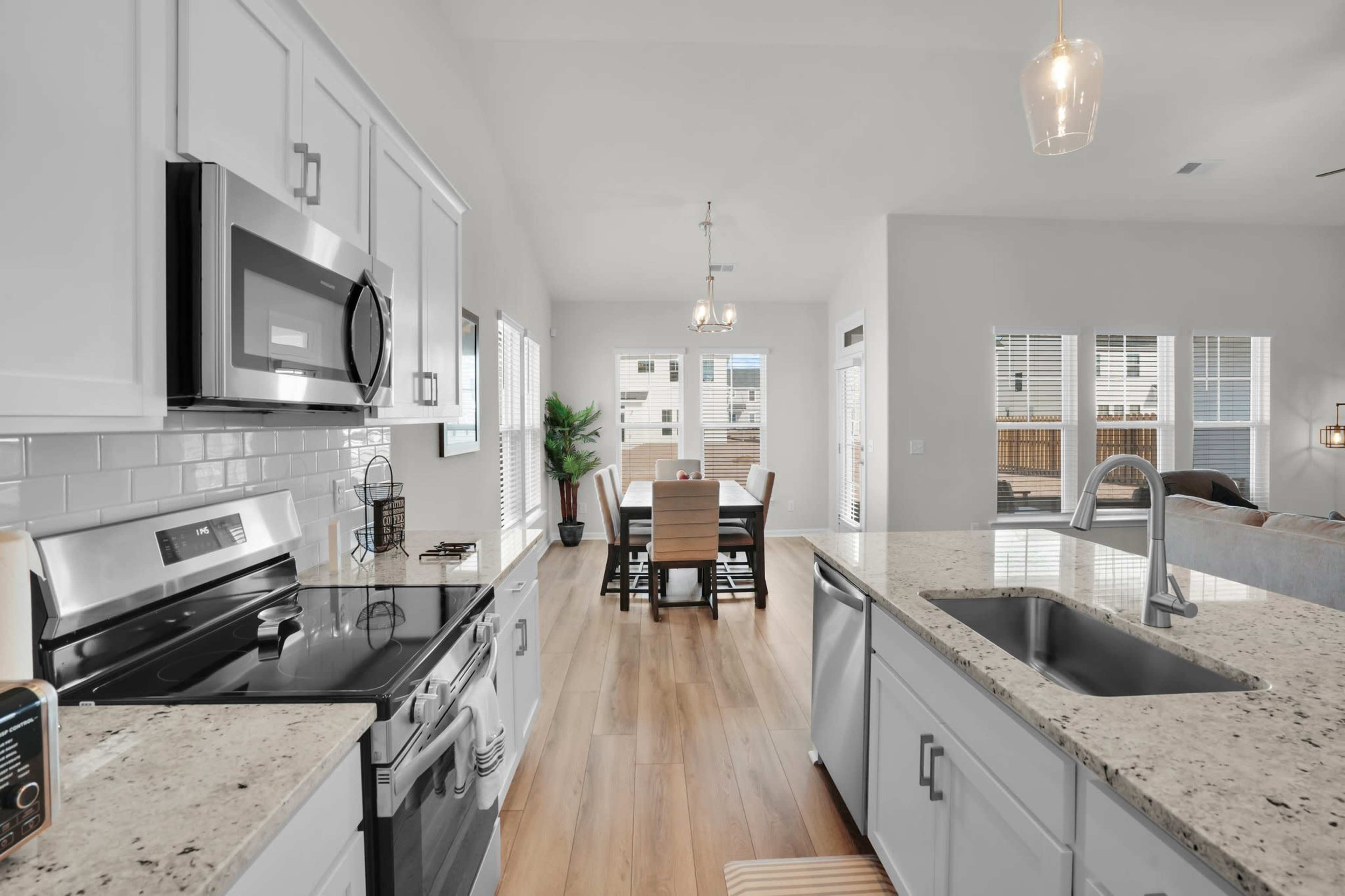 The image shows a modern kitchen with stainless steel appliances, white cabinetry, and a granite countertop that opens up to a dining area and living room.