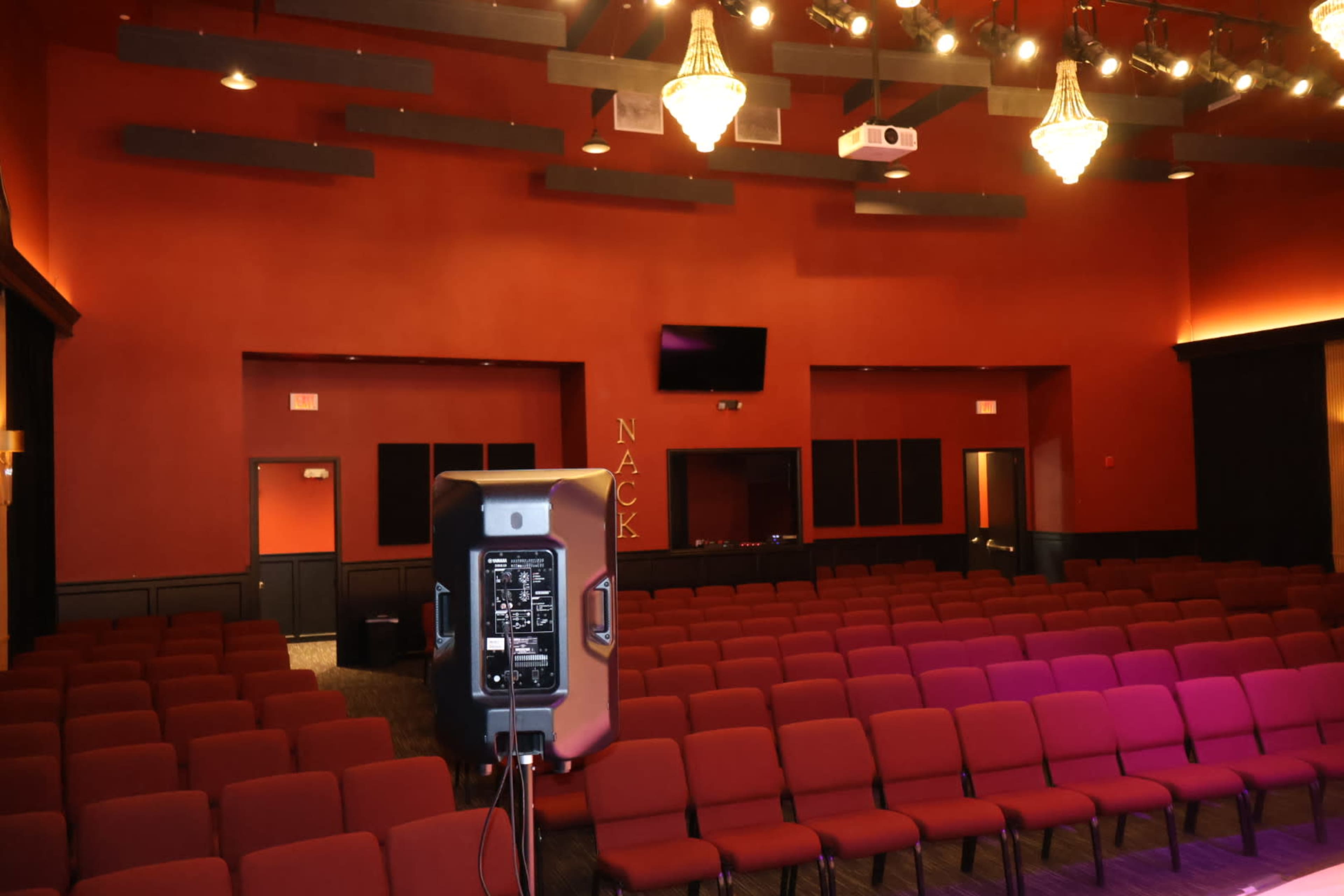 The image shows a red-walled performance space with rows of maroon seats and chandeliers overhead.
