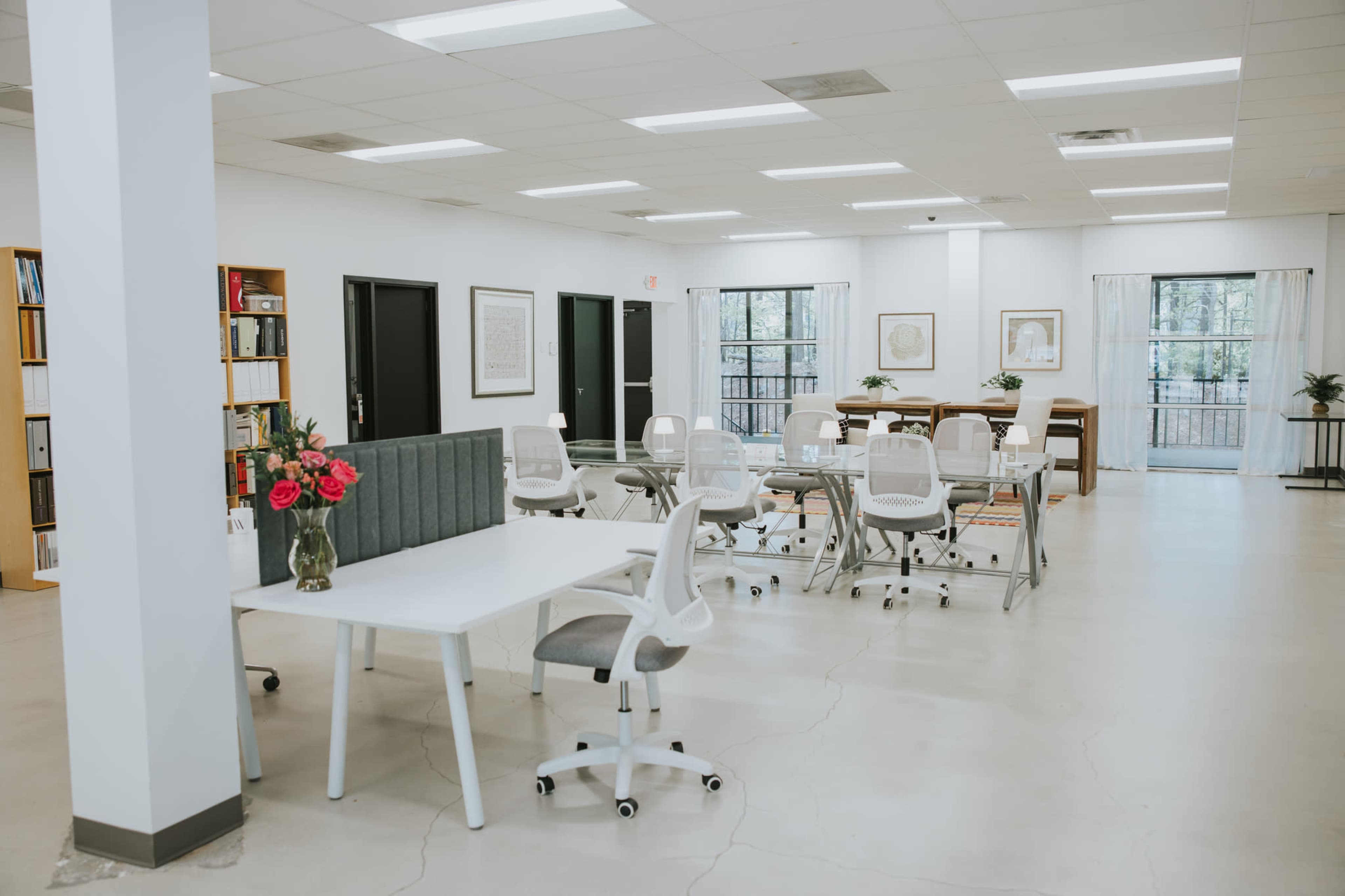 The image shows a spacious office interior with multiple workstations arranged around a central area, featuring white furniture, potted plants, and large windows.