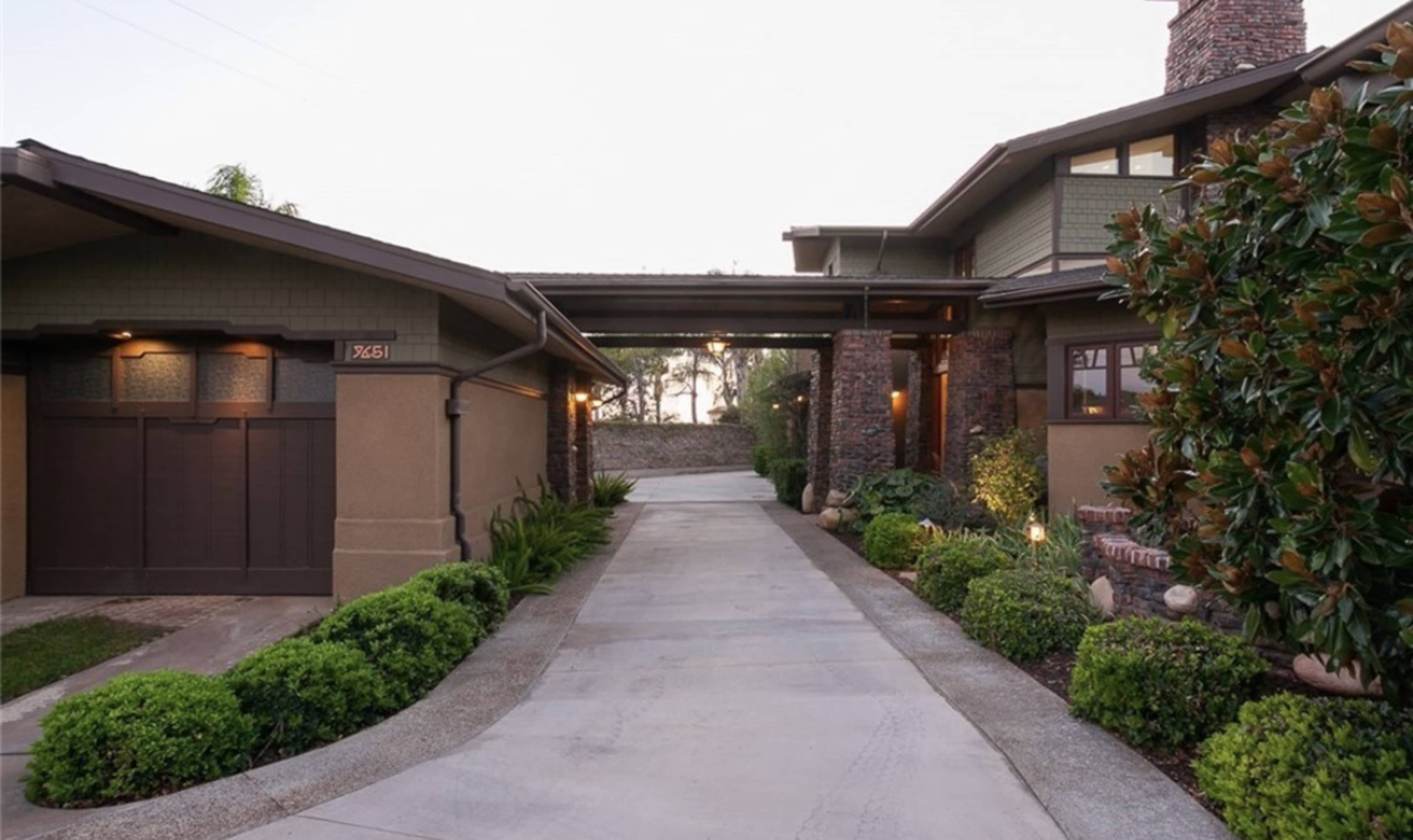 A driveway leads to a two-story house with a covered entrance and landscaped greenery on either side.