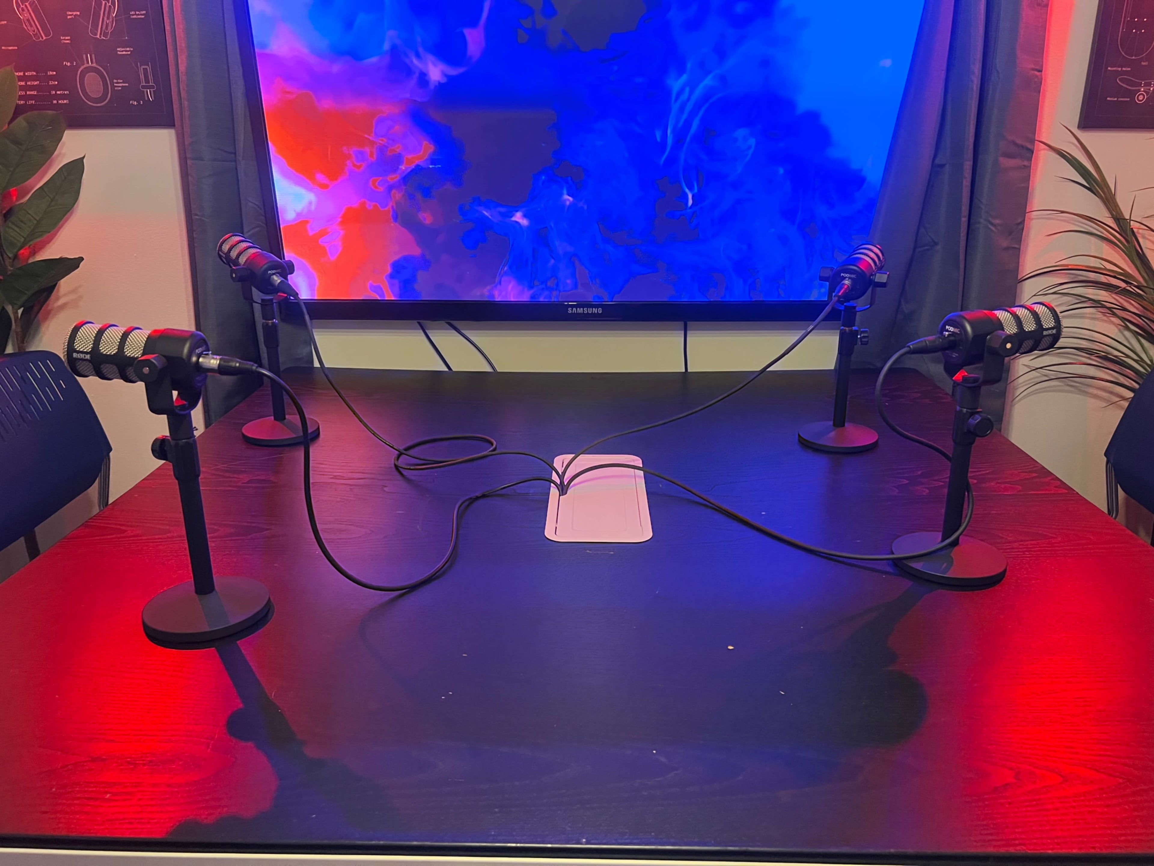 A wooden table with four microphones attached to stands, connected by cables, sits in front of a colorful backdrop on a television screen.