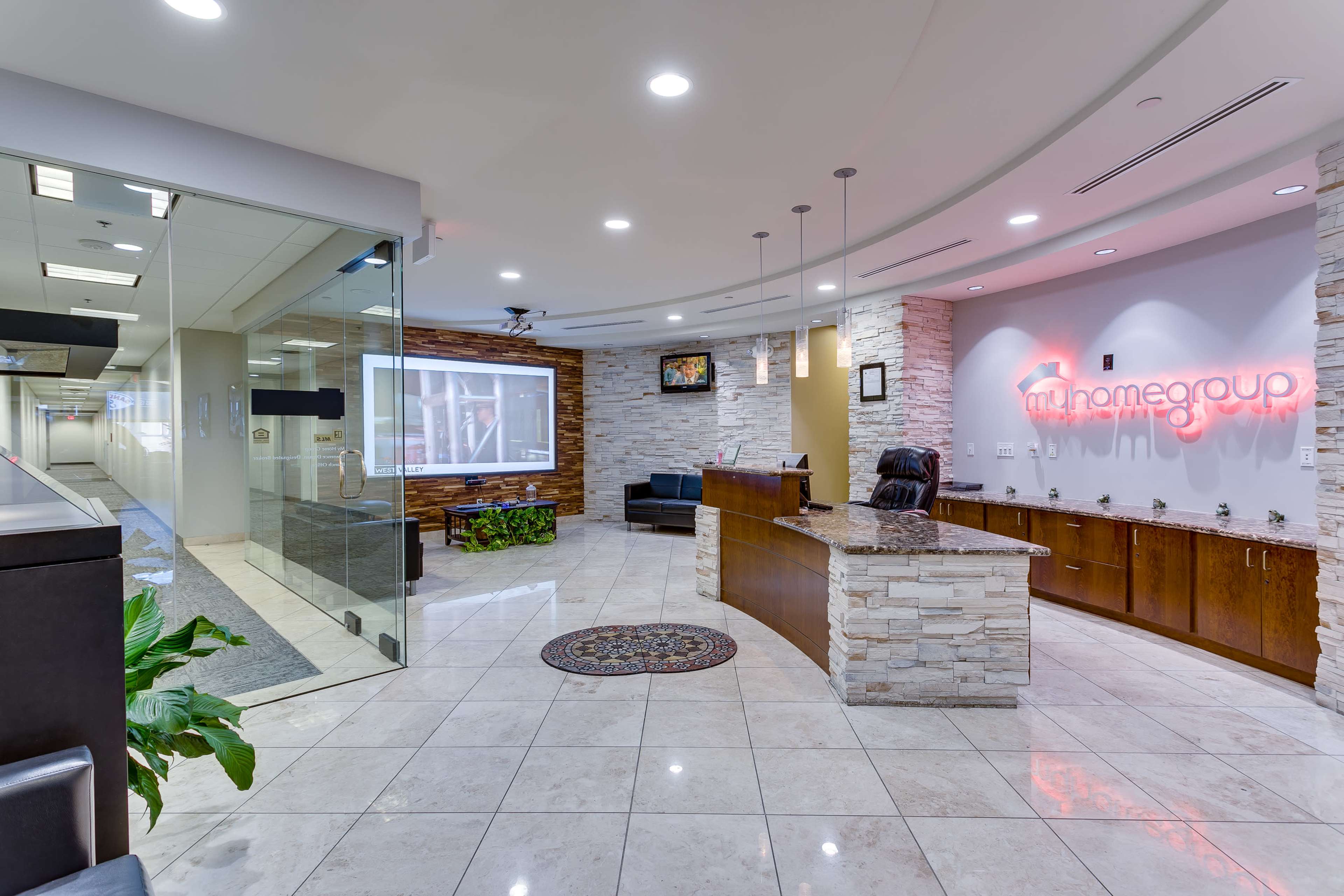 A modern reception area featuring a stone-accented front desk, comfortable seating, and a decorative rug on a tiled floor, with branding displayed on the wall.