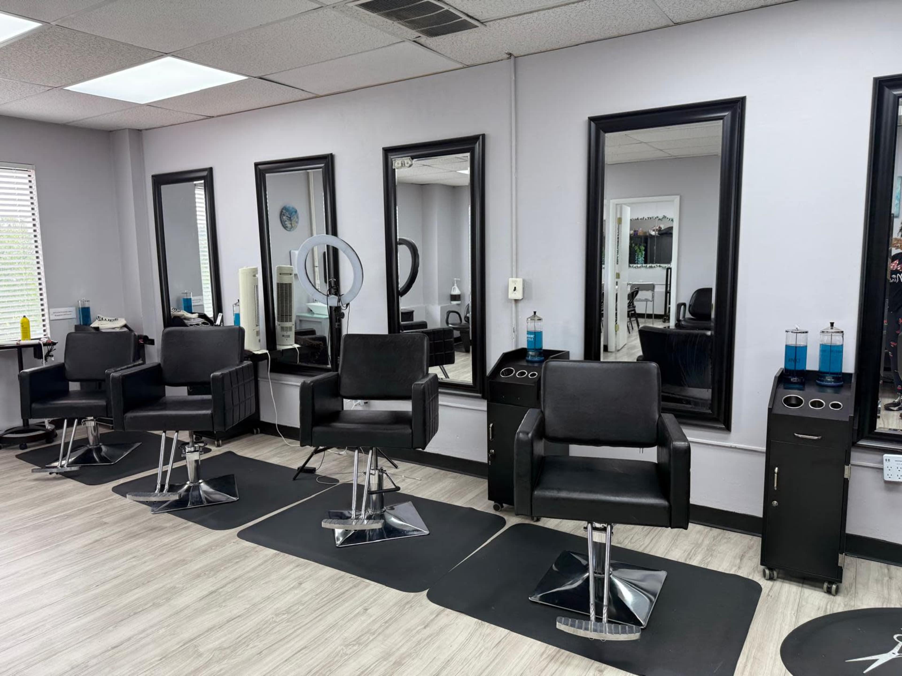 The image shows a modern hair salon with four black styling chairs arranged in front of mirrors, and various styling products placed on nearby counters.