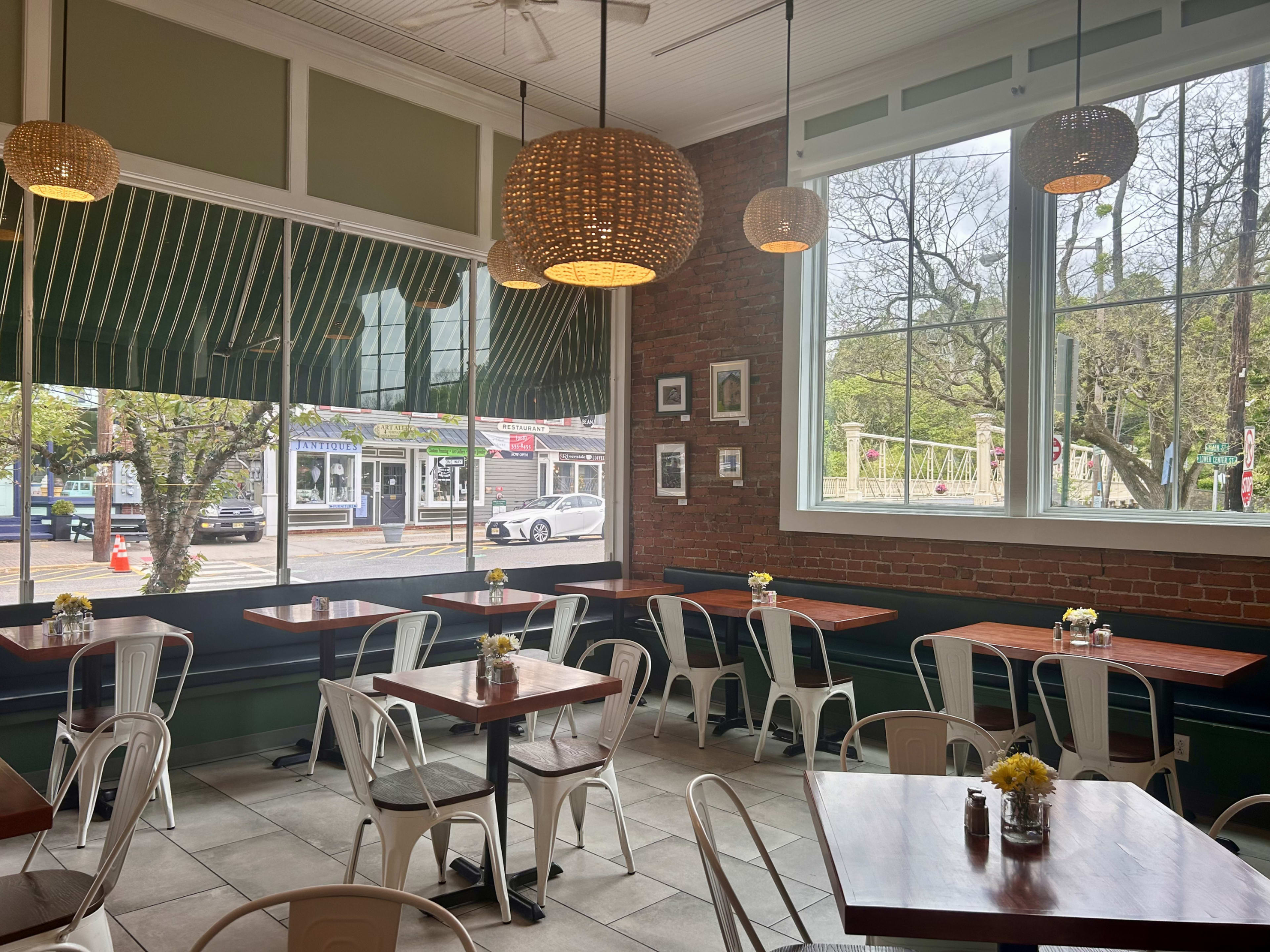 The image shows a cozy café interior featuring wooden tables, metal chairs, and large windows with natural light illuminating the space.