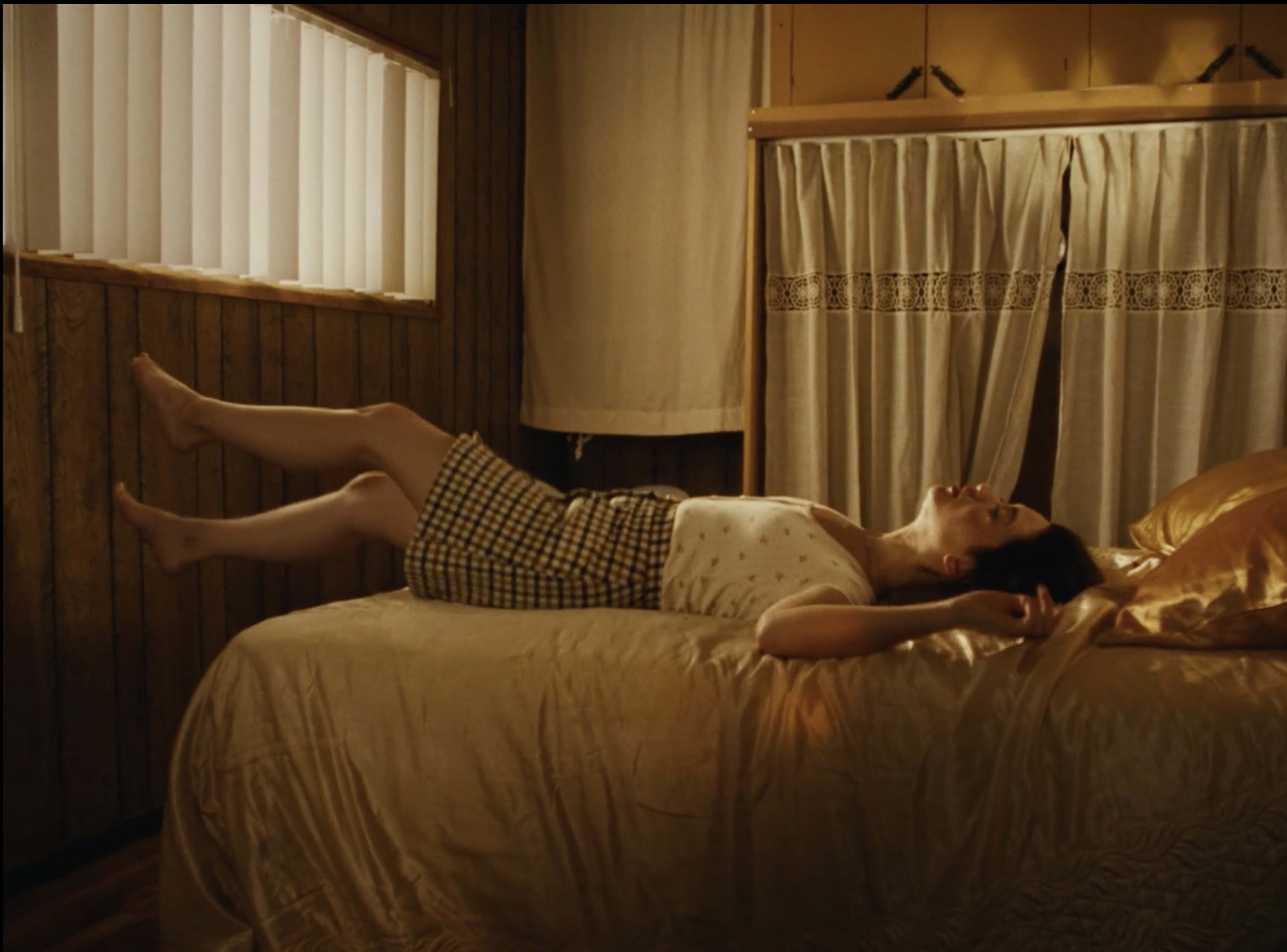 A woman lies on a bed in a wooden room, with her legs raised and eyes closed.