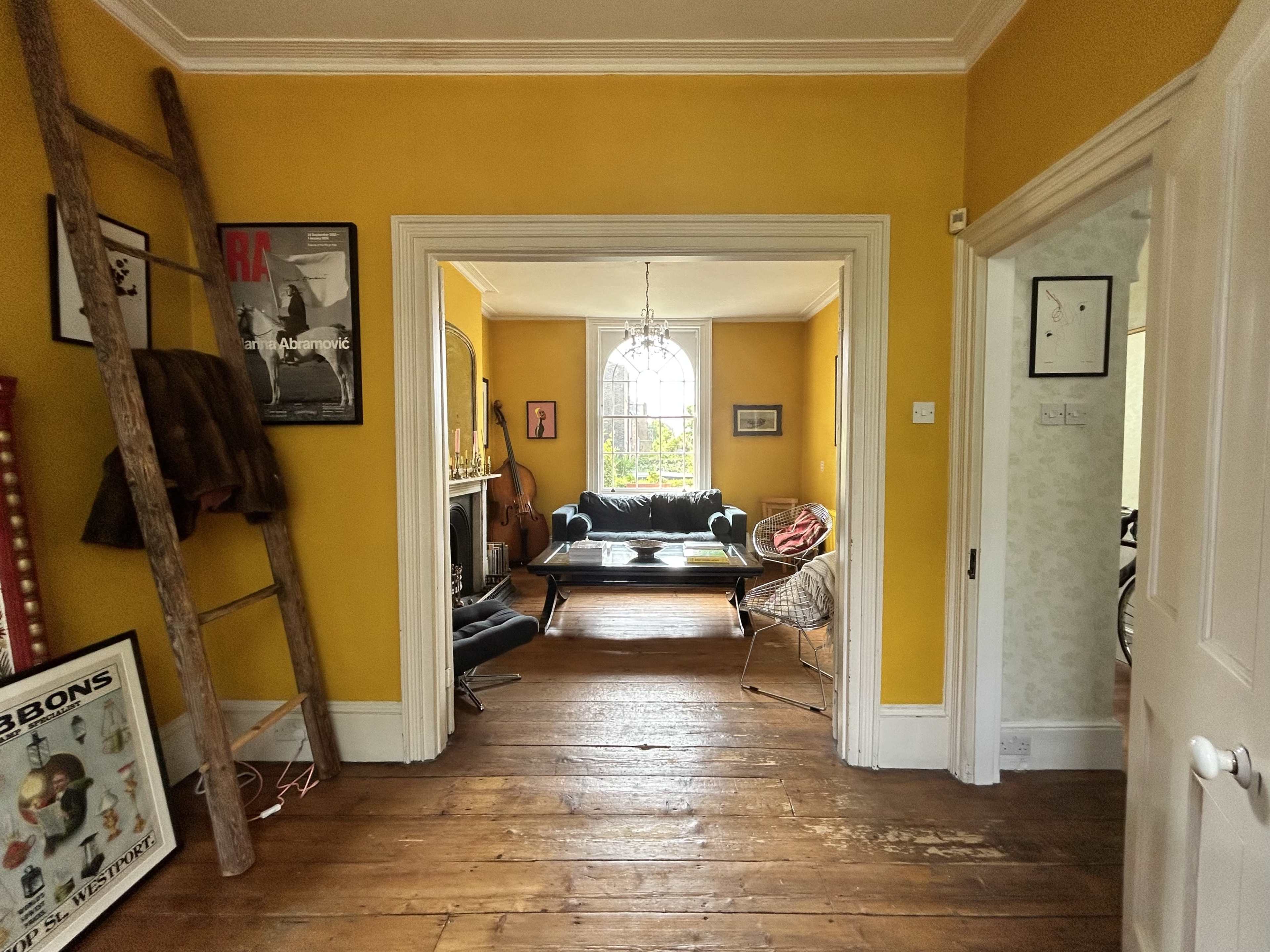 The image depicts a hallway with yellow walls, a wooden ladder, and a view into a bright living room featuring furniture and decorative elements.