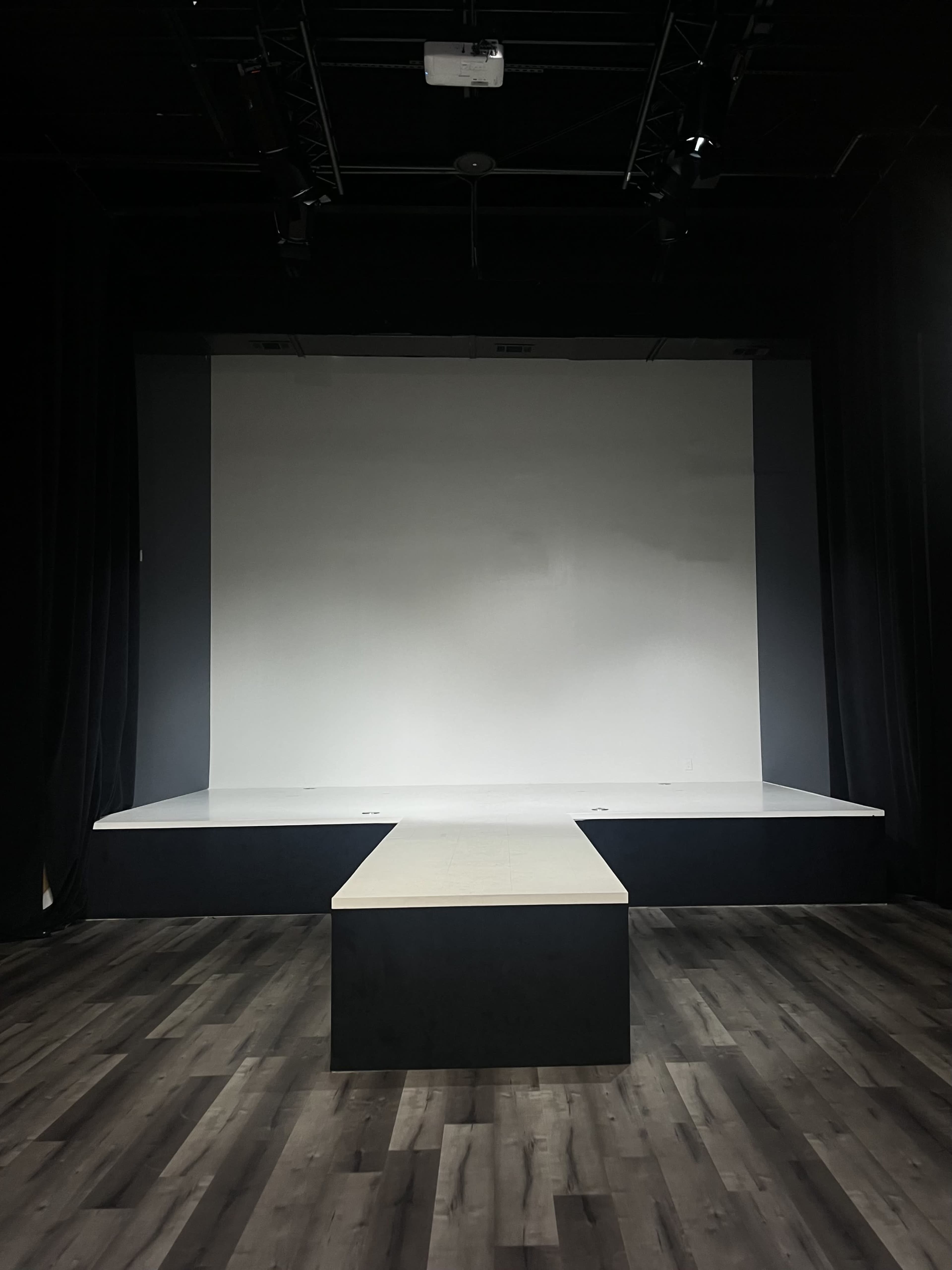 The image shows a minimalist stage with a long, narrow platform extending into a blank white backdrop, surrounded by black curtains.