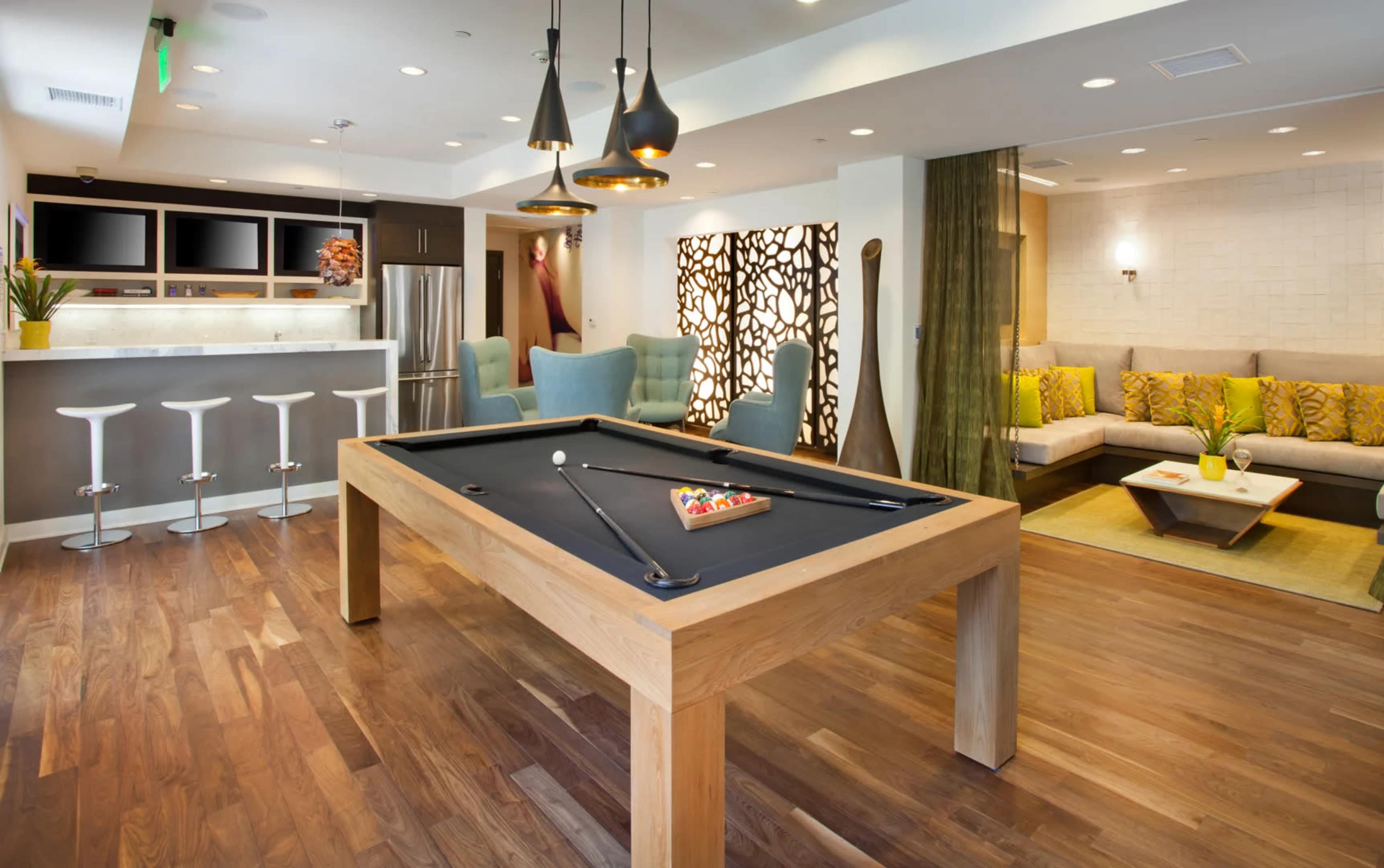 A modern recreational room features a pool table, seating area with a sofa and chairs, and a kitchenette in the background.