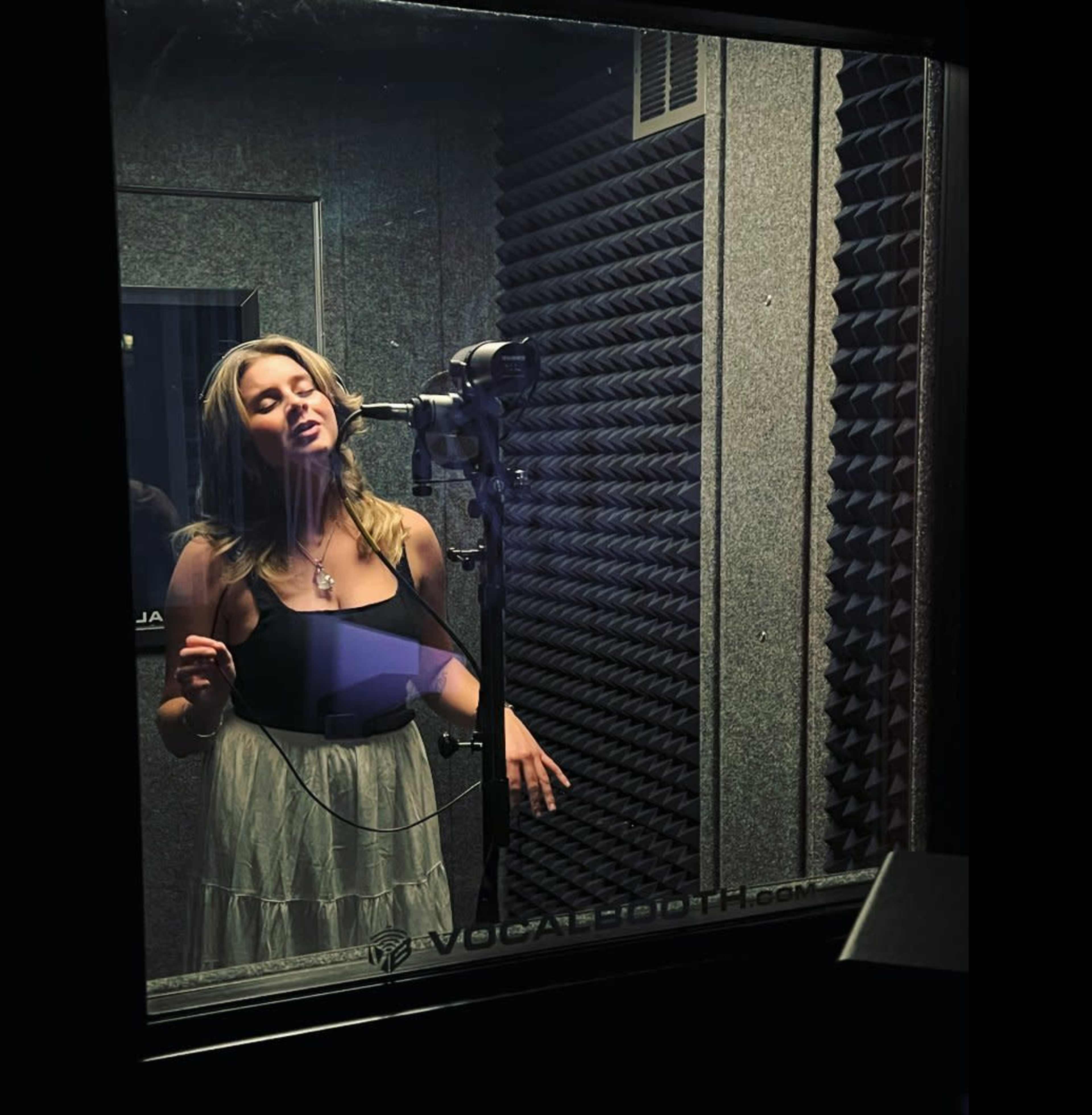 A woman stands in a soundproof recording booth, singing into a microphone.