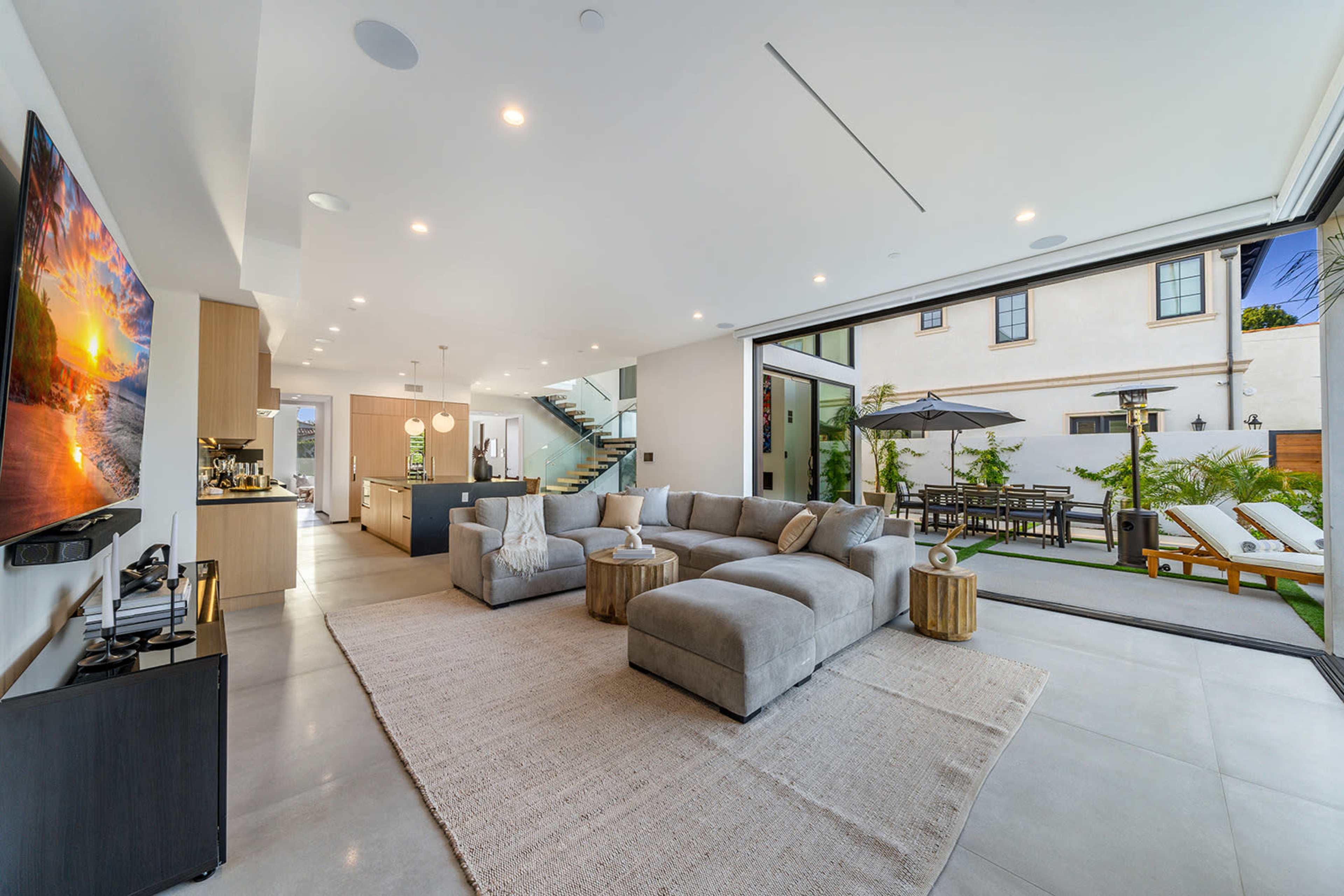 A modern living room with large sliding doors opening to a patio and garden area, featuring a sectional sofa, a coffee table, and a flat-screen TV mounted on the wall.