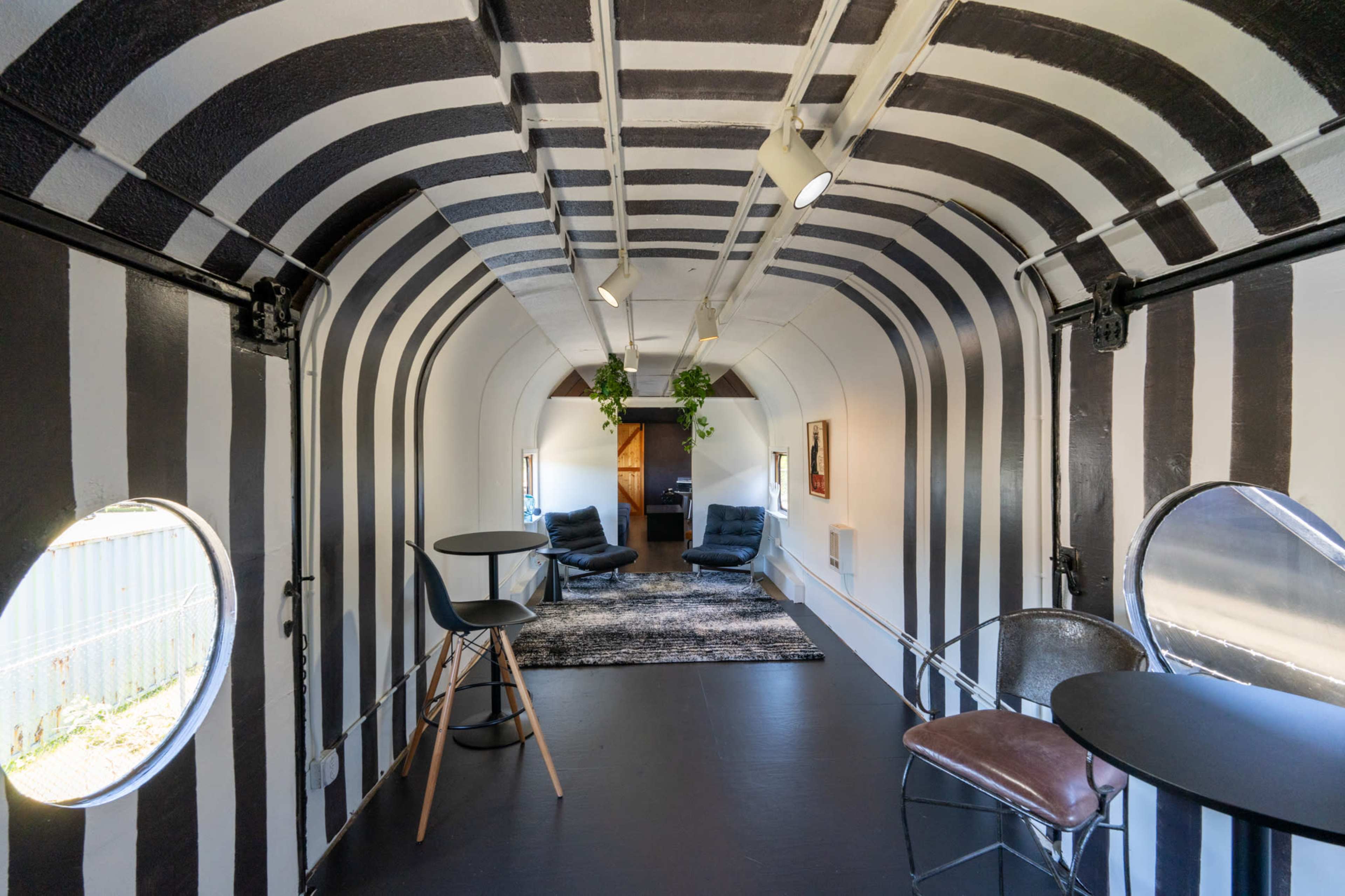 The image shows a narrow, tunnel-like room featuring black and white striped walls, a plush rug, and two seating areas with chairs and tables at either end.