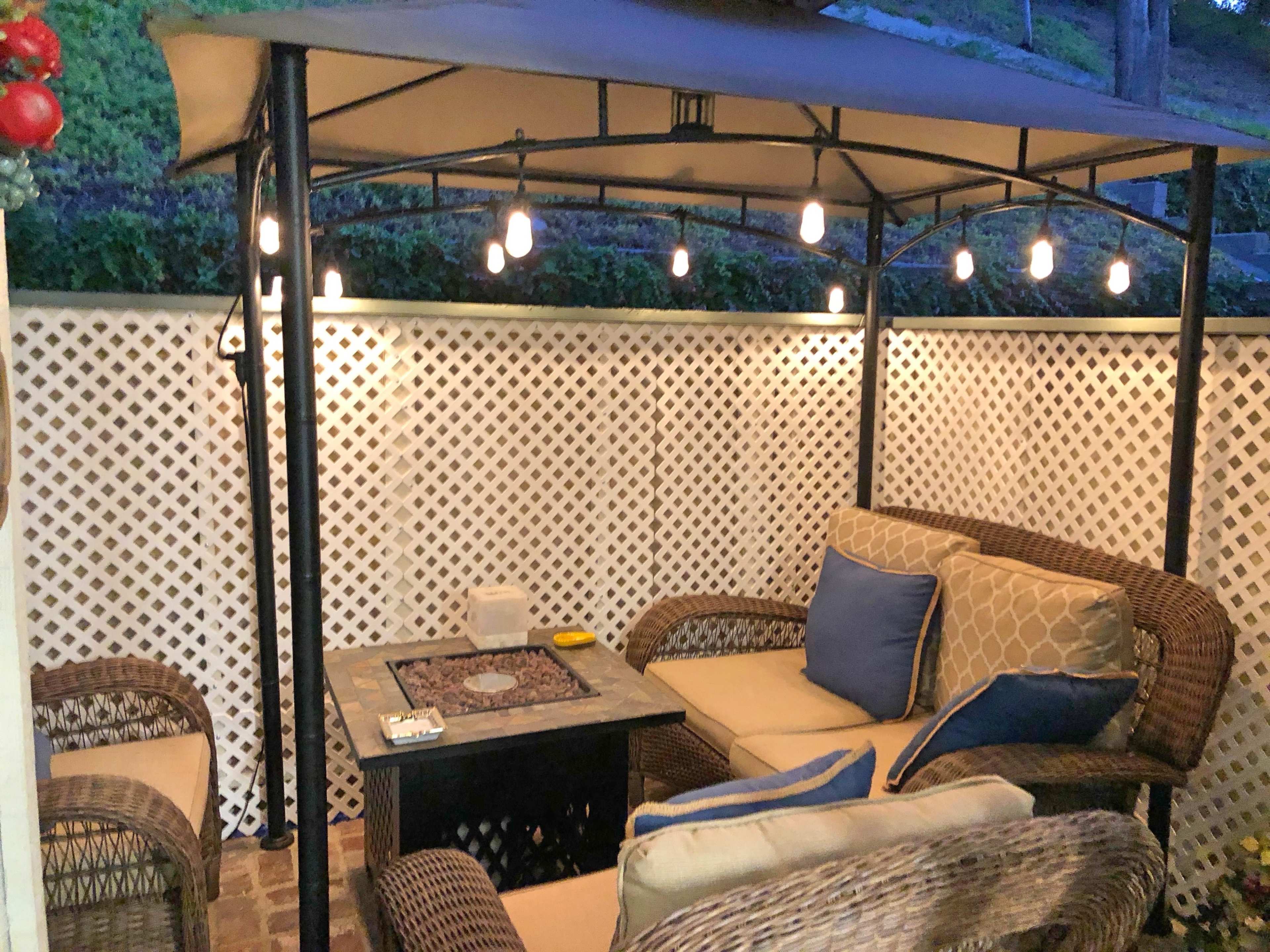 The scene shows a cozy outdoor seating area with a canopy, illuminated by string lights, featuring wicker furniture and a small table.