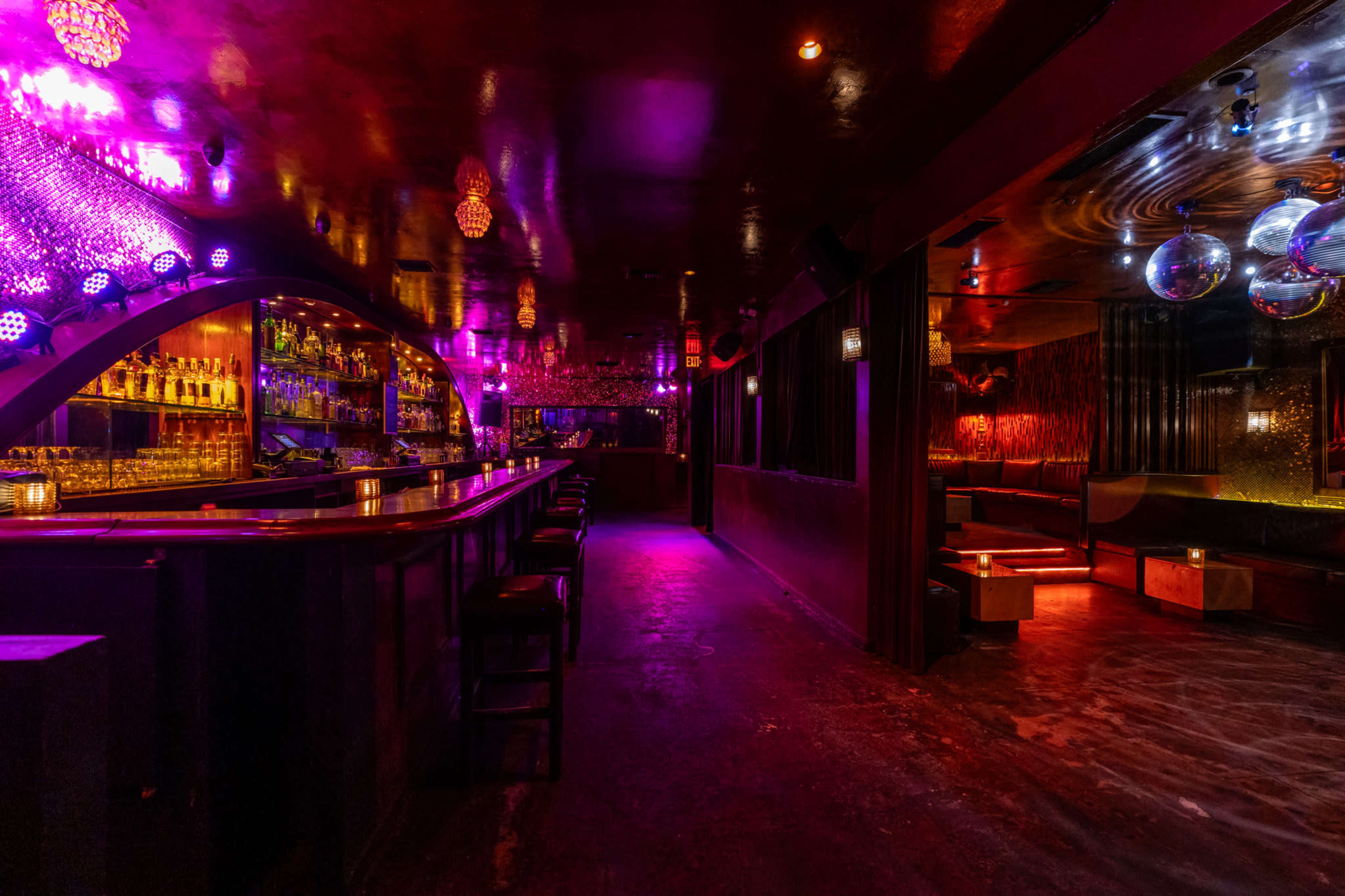 A dimly lit bar interior featuring a long bar counter, illuminated shelves of alcohol, and colorful light fixtures reflecting off the walls.