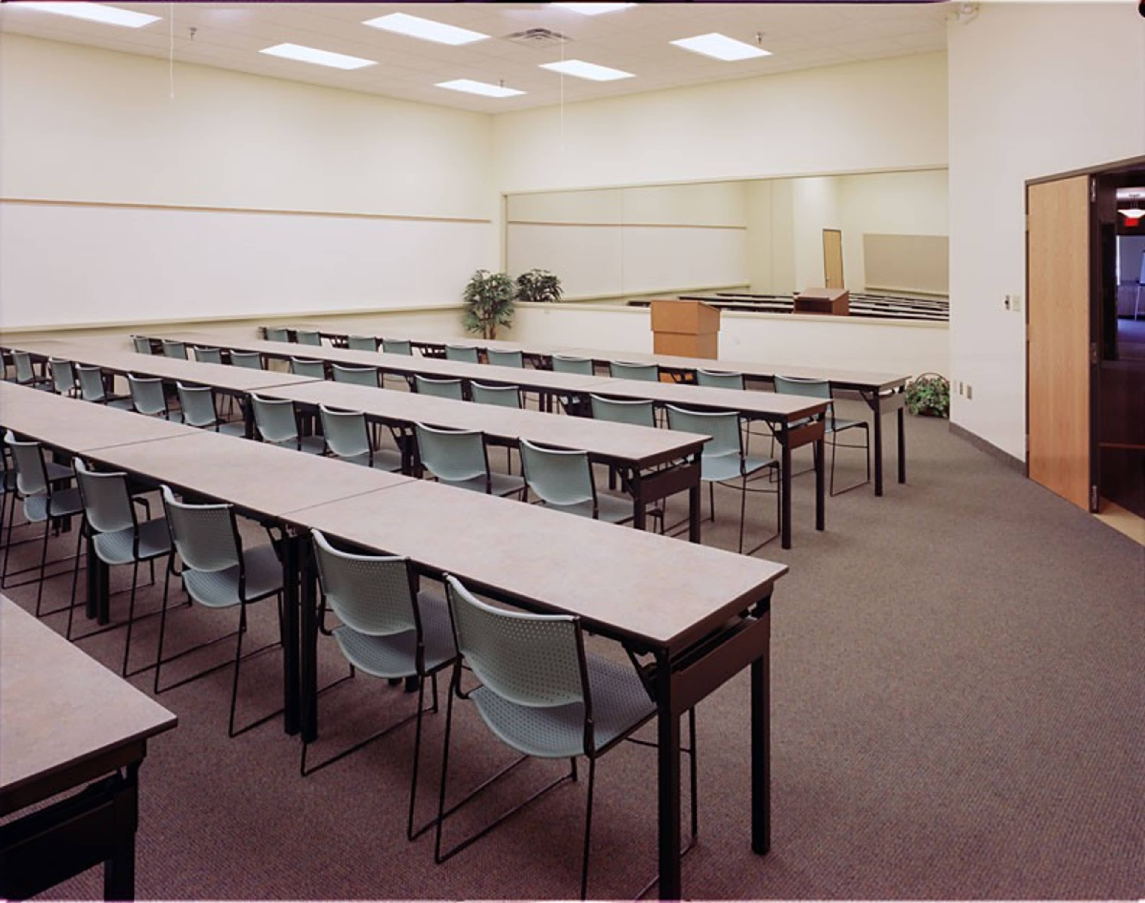 A classroom is set up with rows of tables and chairs facing a wooden podium and a mirrored wall.