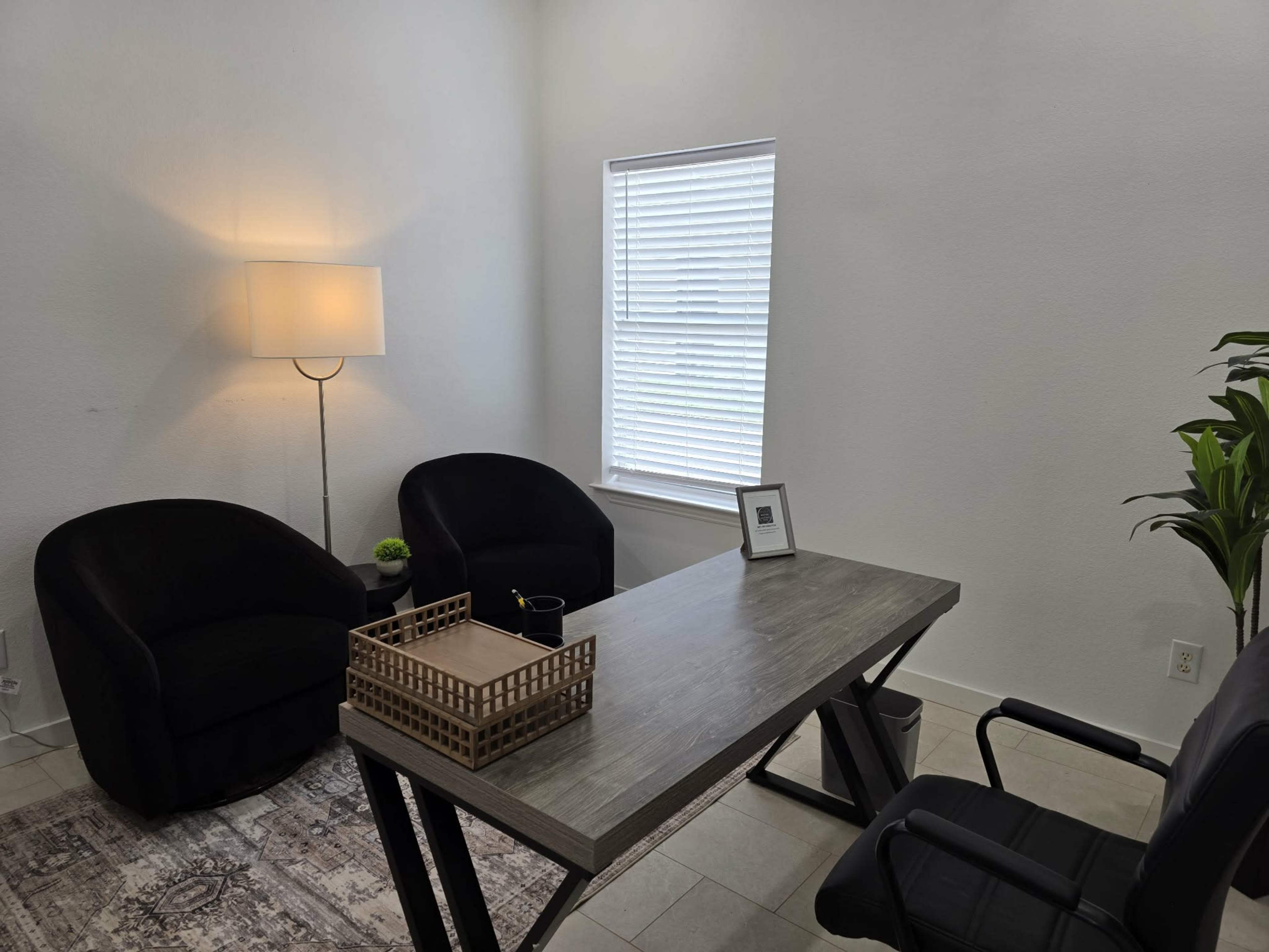 The image shows a well-lit office space featuring a desk, two black armchairs, a decorative tray on the desk, and a potted plant near the window.