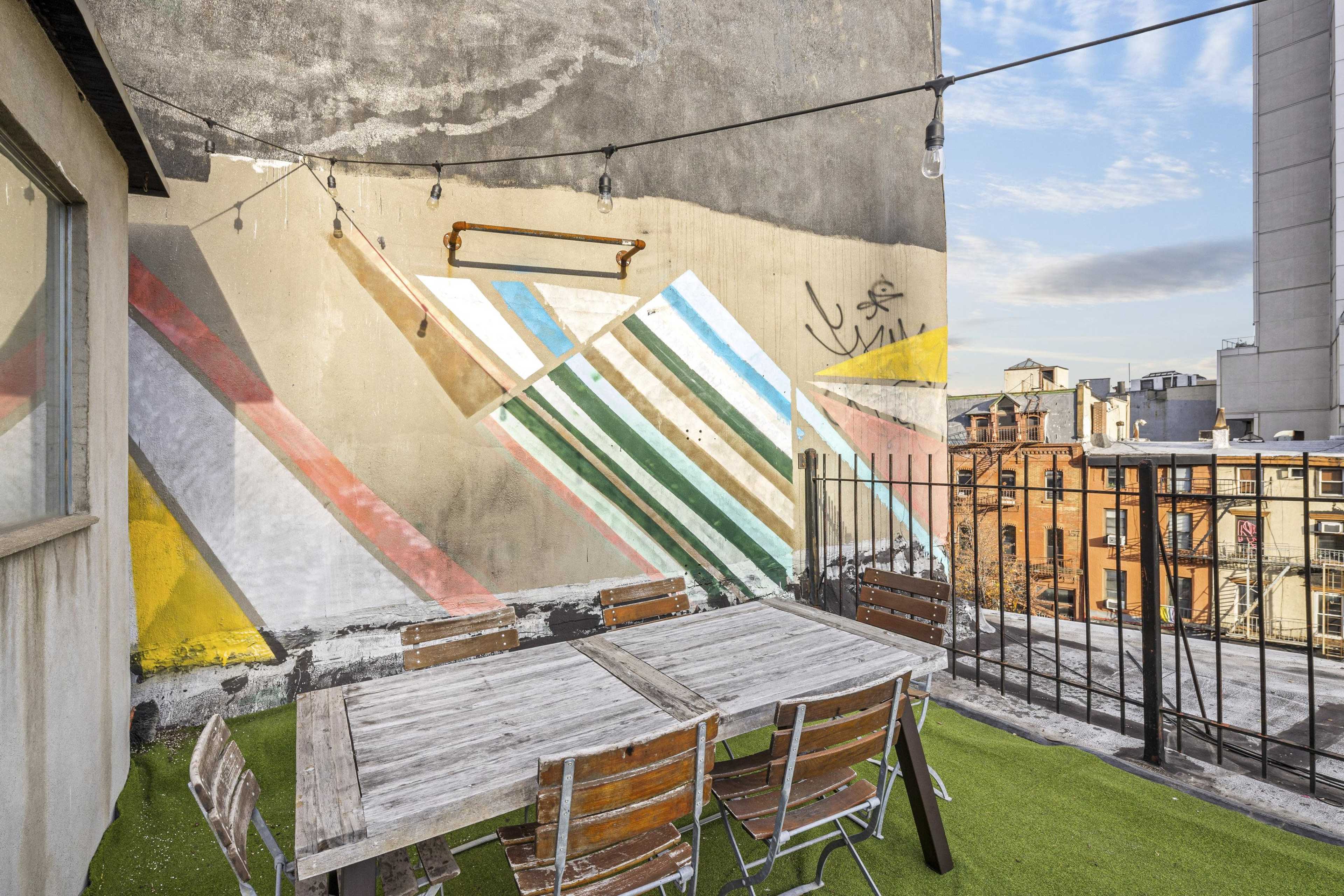 A rooftop terrace features a wooden table surrounded by metal chairs, with a colorful mural on the wall and string lights above.