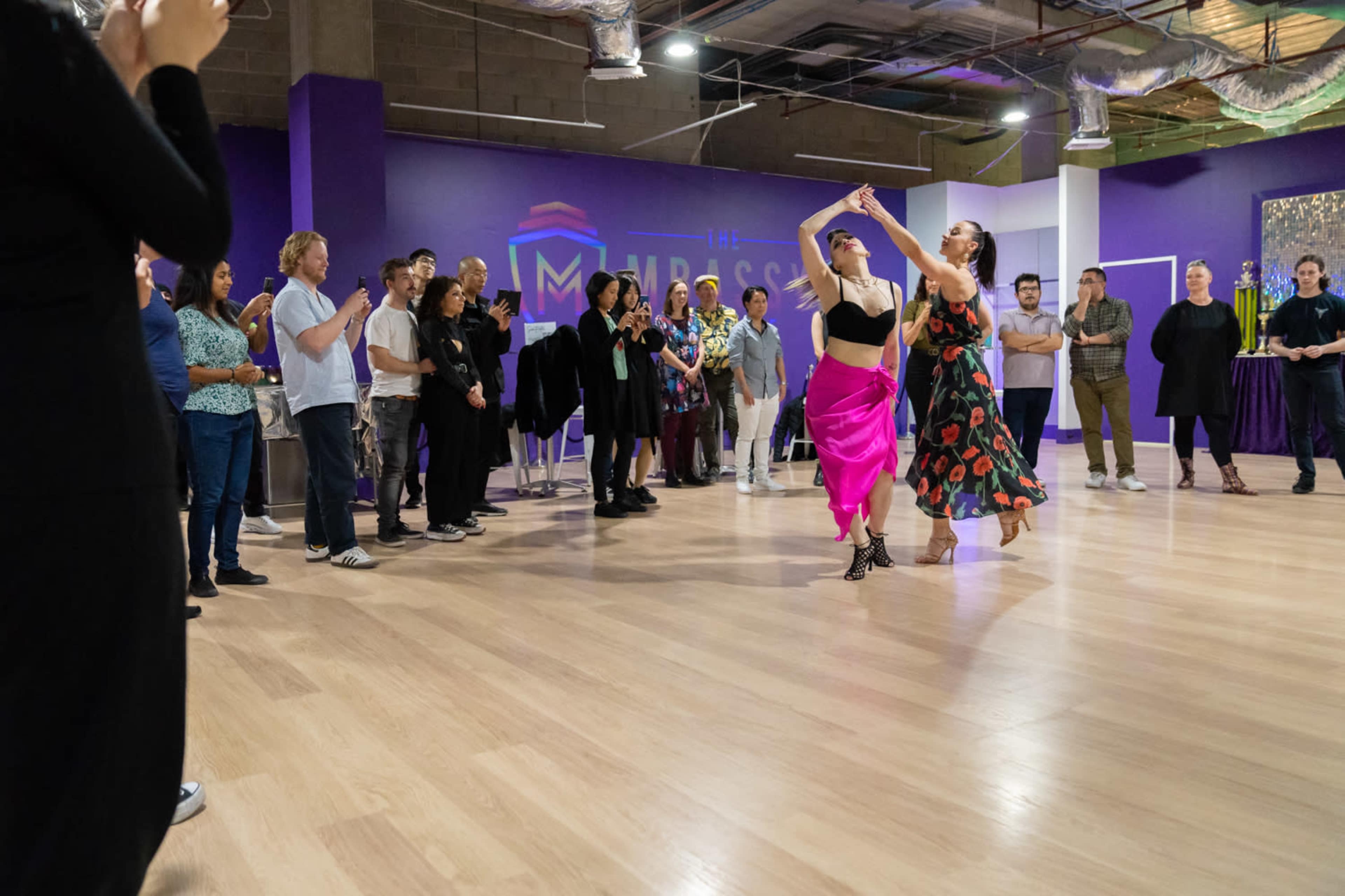 Two dancers perform in a brightly lit studio while an audience of onlookers watches and applauds.