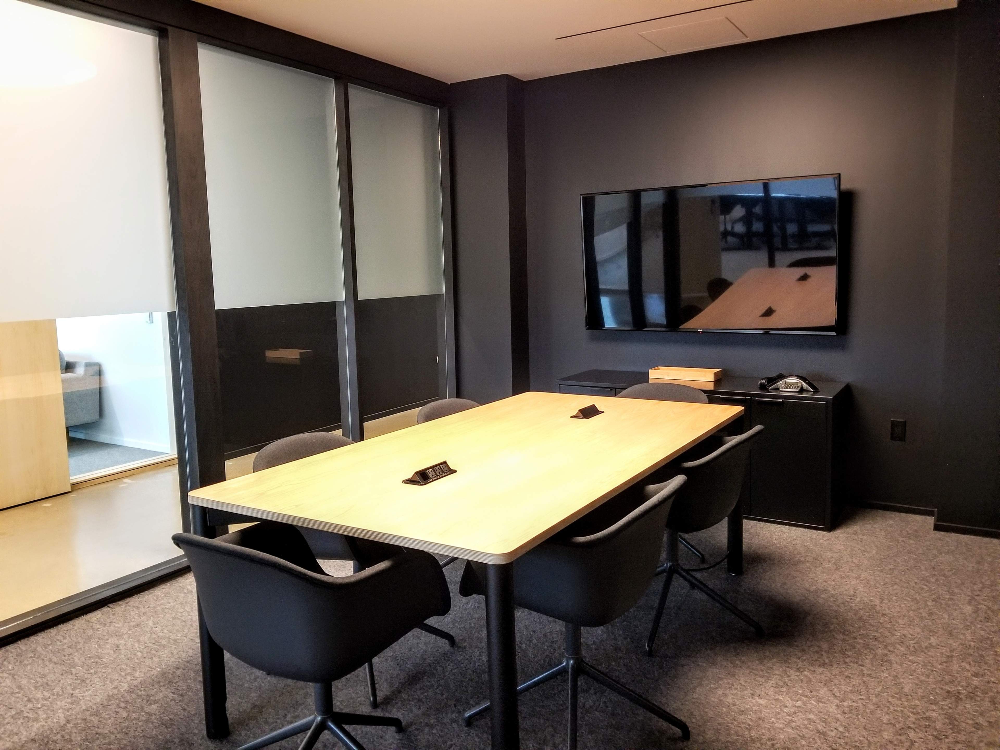 A modern conference room featuring a wooden table surrounded by four chairs, a large television on the wall, and glass partitions leading to another area.