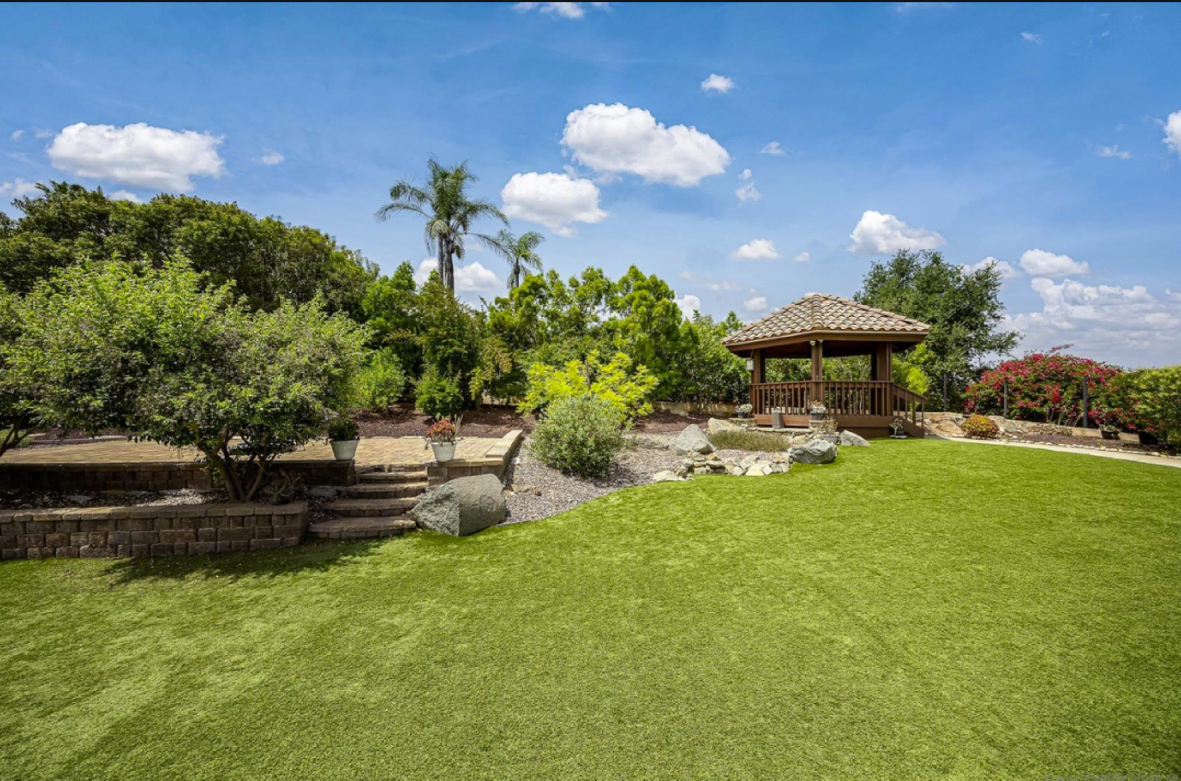 The image shows a well-maintained garden with a gazebo, landscaped greenery, and stone pathways under a blue sky with fluffy clouds.