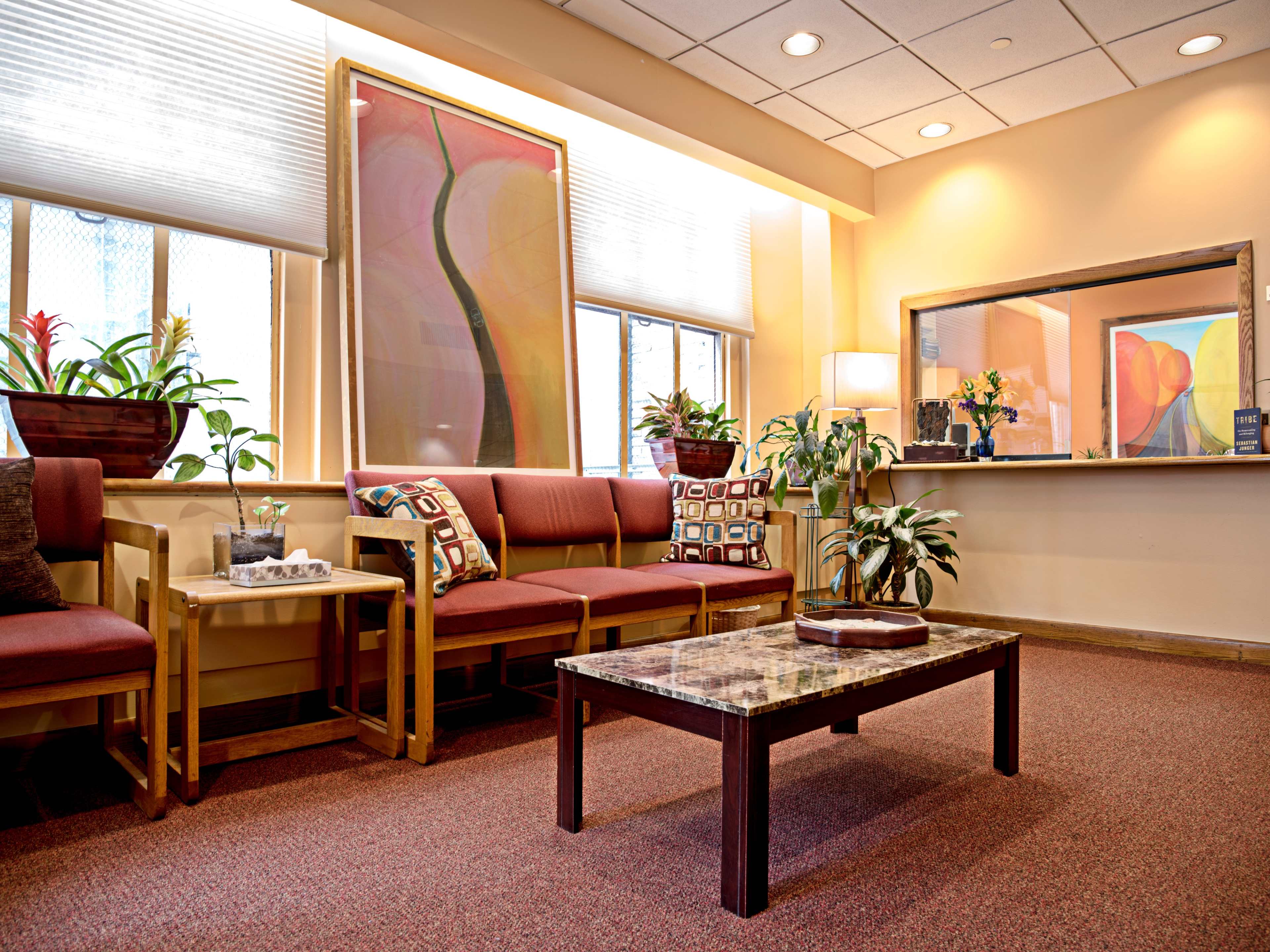 The image displays a waiting area with red upholstery seating, potted plants, and a coffee table in front of a mirror and colorful artwork on the walls.